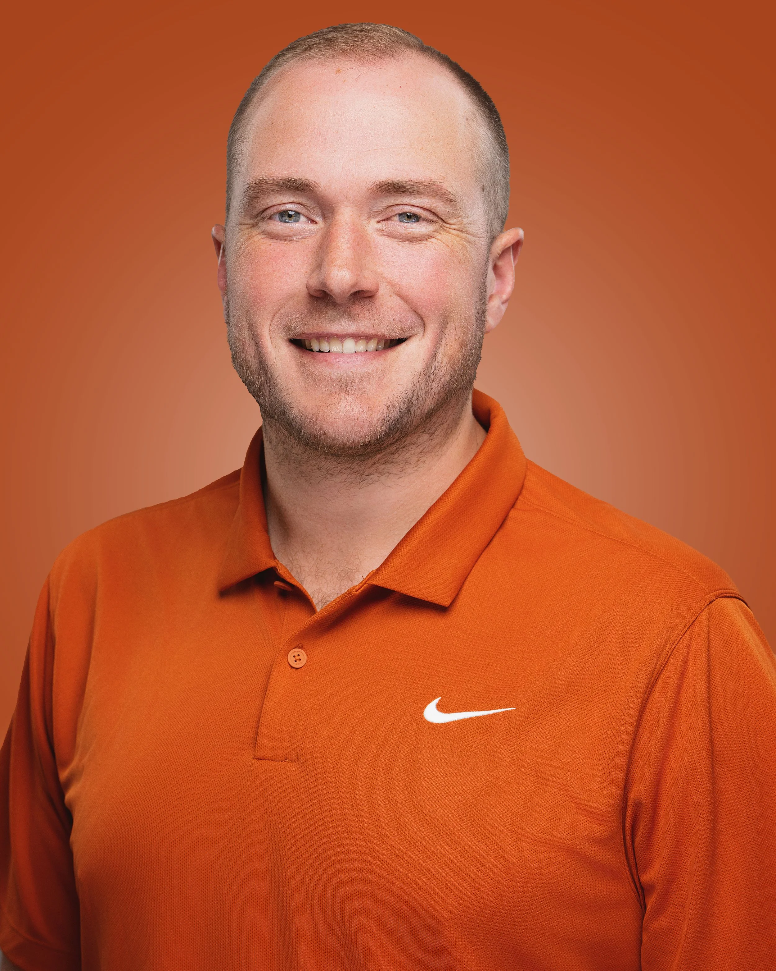 A smiling man wearing an orange Nike polo shirt against an orange background.