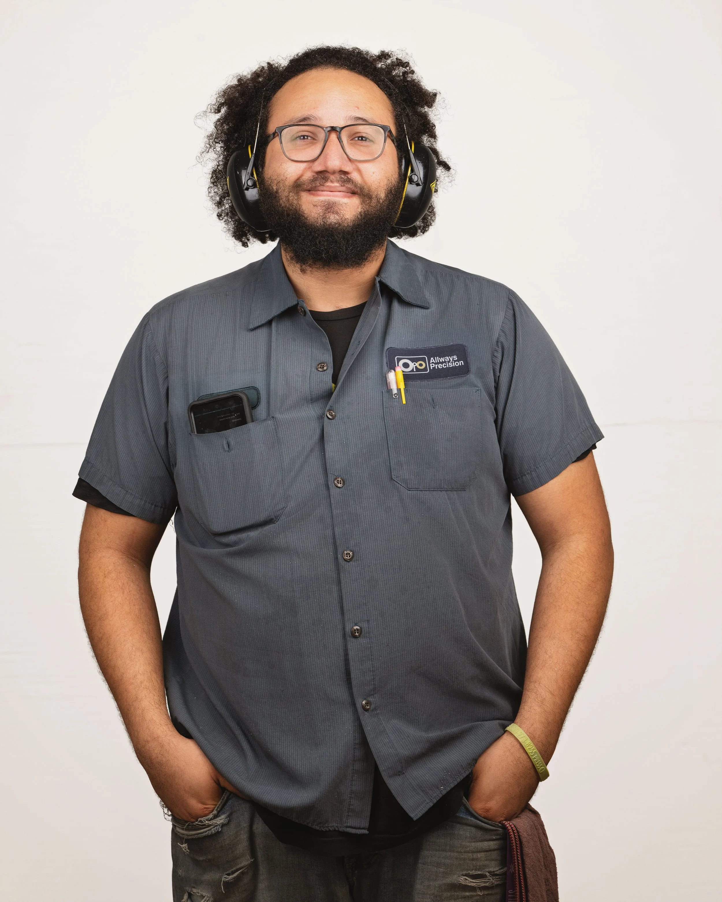 A man with curly hair and a beard wearing glasses, a gray work shirt with a logo that says 'Allways Precision,' and headphones, standing against a plain white background.