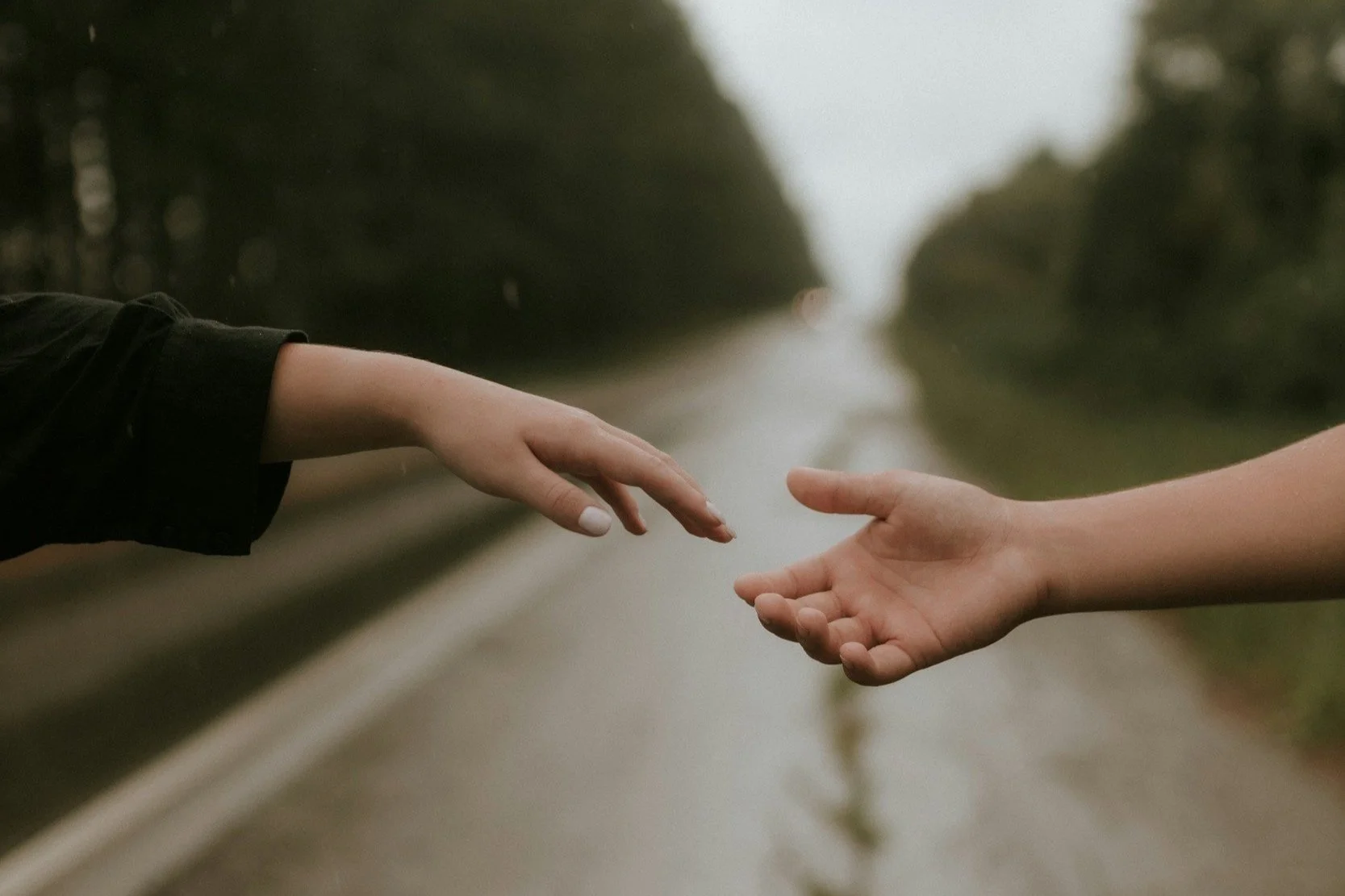 Two hands reaching toward each other over an empty road with trees on both sides, in a natural outdoor setting.