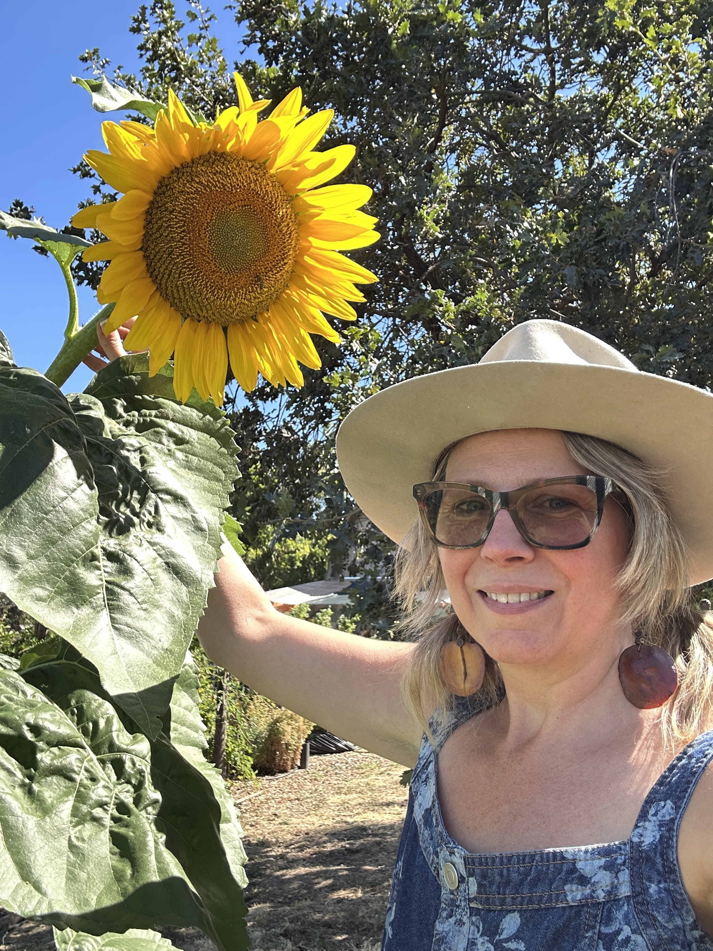 A woman with blonde hair, wearing glasses, a wide-brimmed hat, and earrings, is taking a selfie outdoors. She is smiling and standing next to a large sunflower with yellow petals and a brown center.