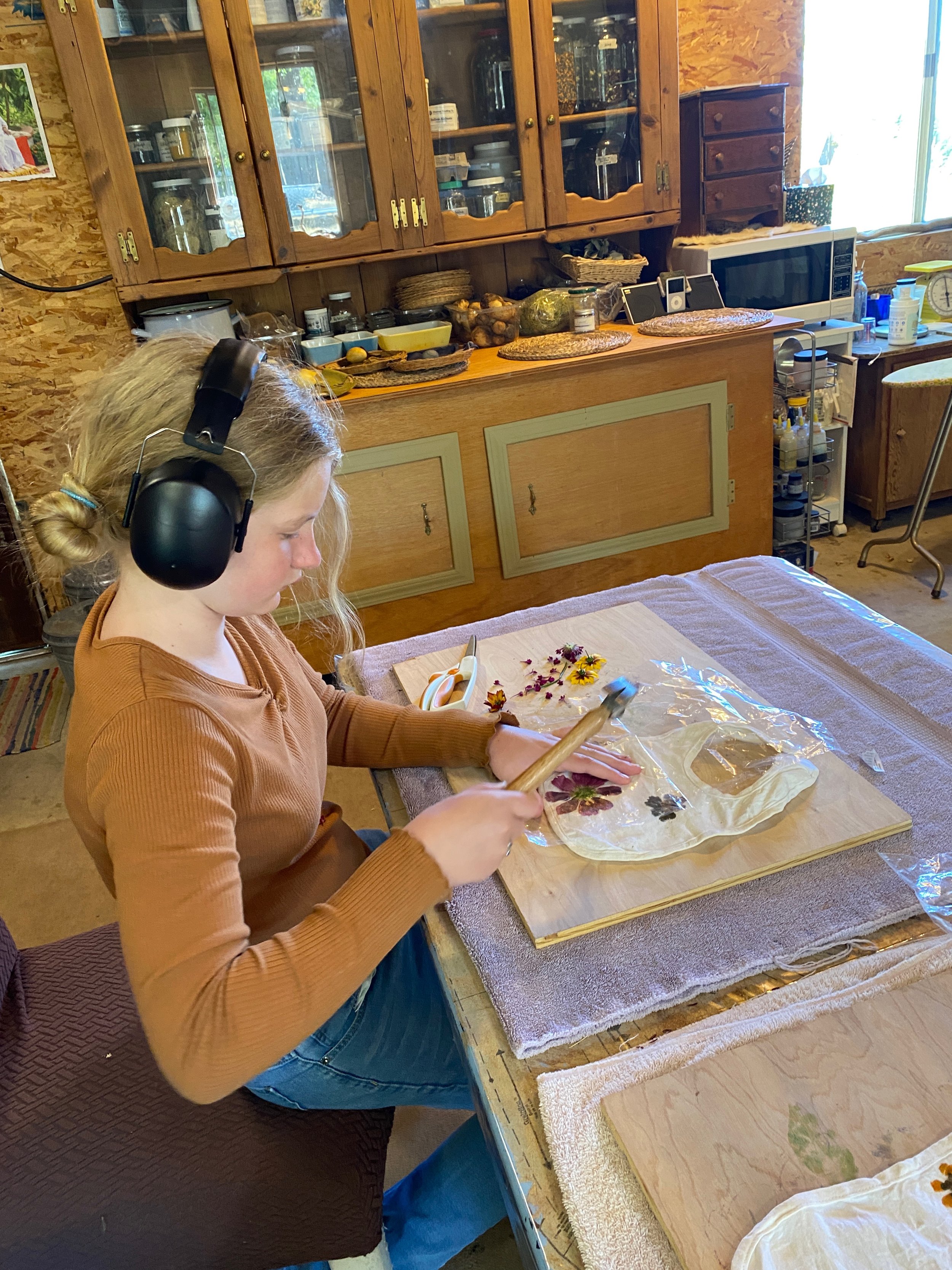 A young woman with blonde hair wearing a brown long-sleeve top and blue jeans is sitting at a wooden table creating pressed flower art. She is wearing large black headphones and is working with pressed flowers on a sheet of plastic or glass, with scissors nearby. Behind her, there is a kitchen with wooden cabinets, jars, bowls, a microwave, and a small drawer unit, on a rustic wood-paneled wall.