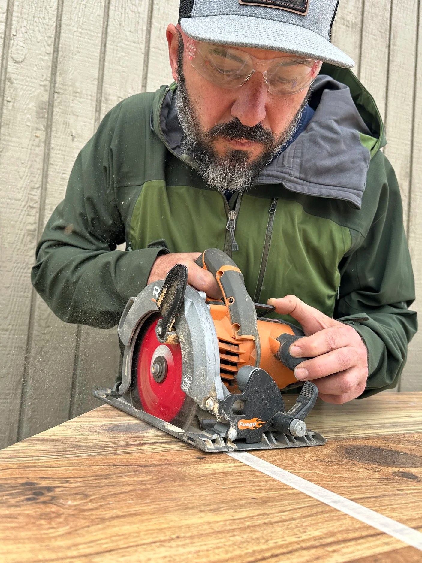 A man with glasses and a beard cutting wood with a circular saw on a wooden table, against a wood-paneled wall background.