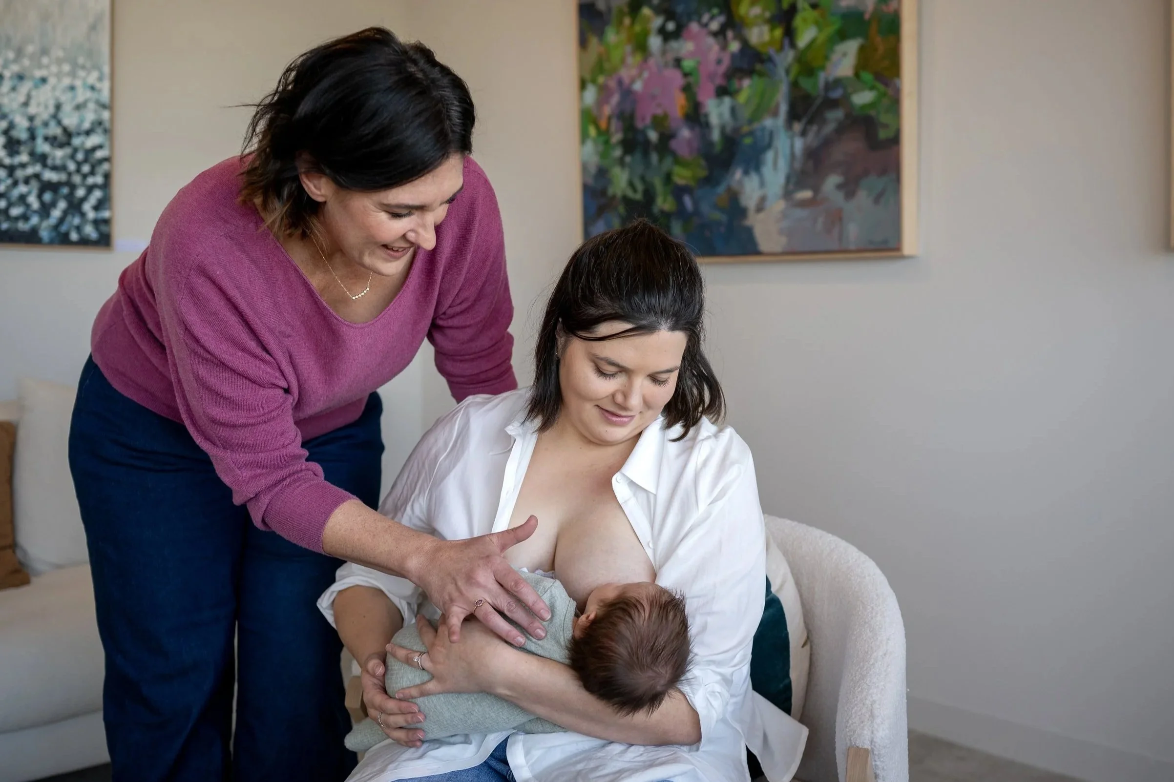 Woman with dark hair holding a newborn baby, smiling and sitting on a white chair. Another woman with short dark hair, wearing a pink sweater, leaning over and touching the baby's hand. The scene is indoors with abstract paintings on the wall.
