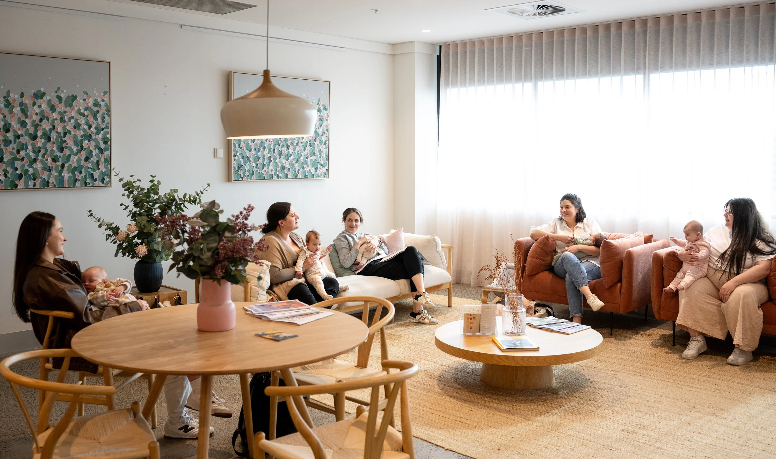 Group of women and babies sitting and chatting in a bright, modern living room with artwork, flowers, and natural sunlight.