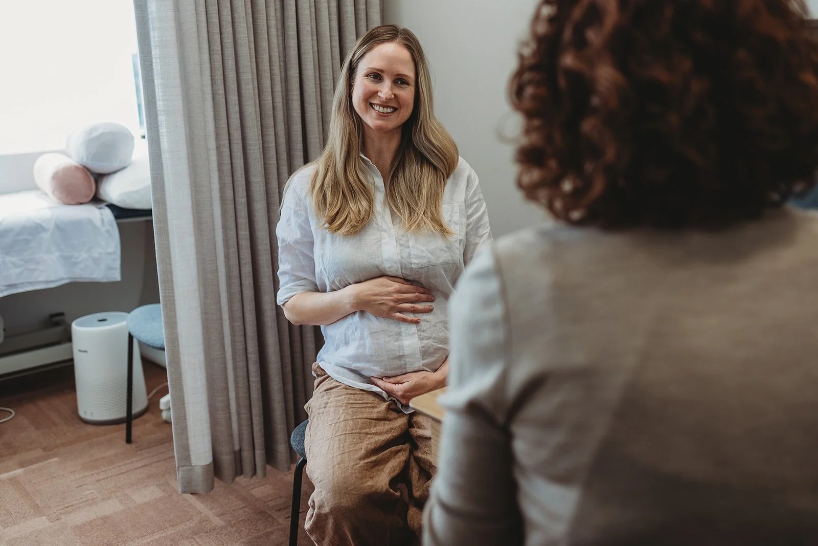 A pregnant woman smiling and holding her belly while talking to another woman in a consultation room.