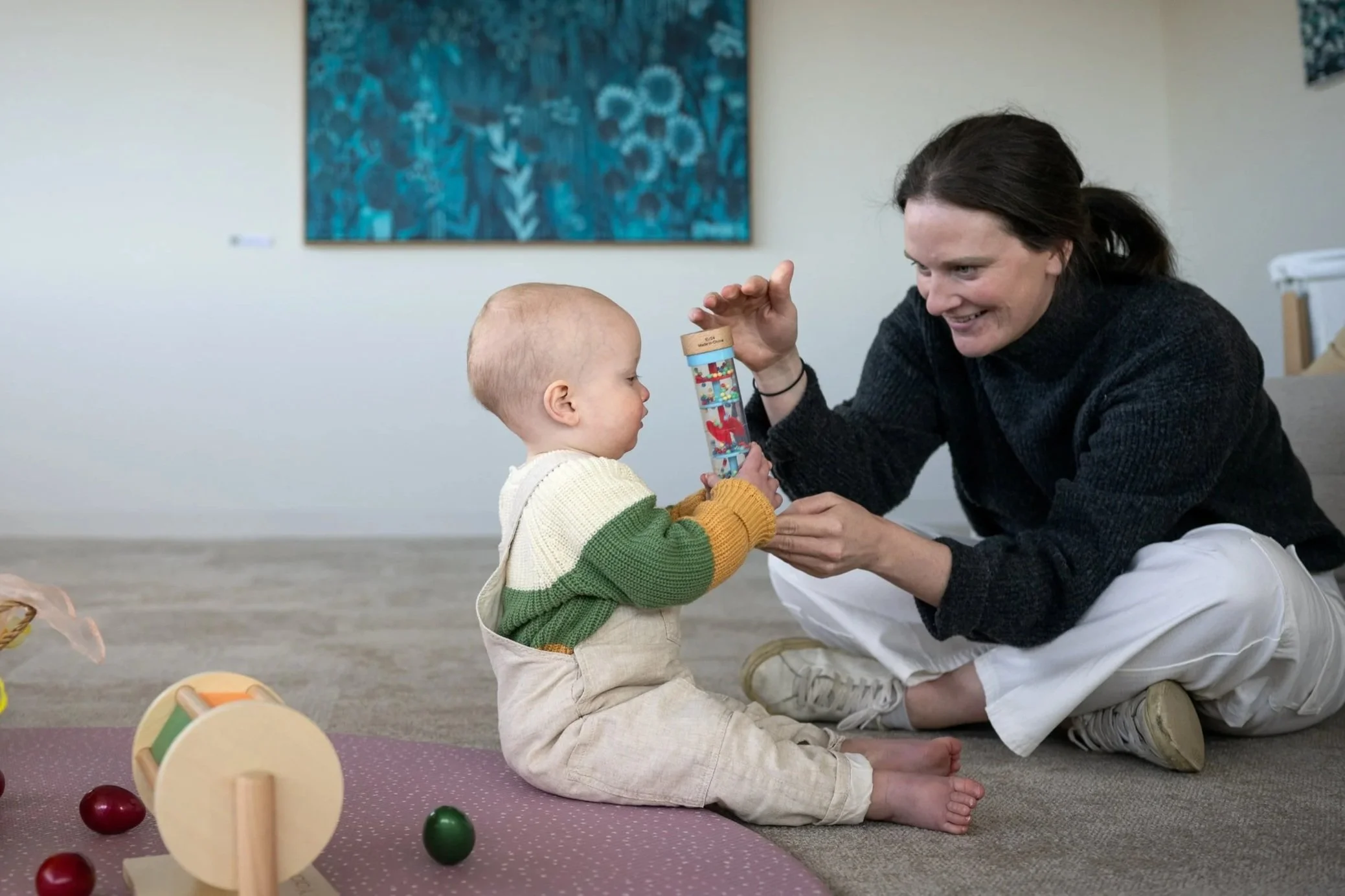 A woman and a young child sit on the floor playing with a wooden toy and a tube with small colorful objects inside as part of Maternal and Infant Wellbeing Melbourne's Sensory Class with Andrea Hunt.