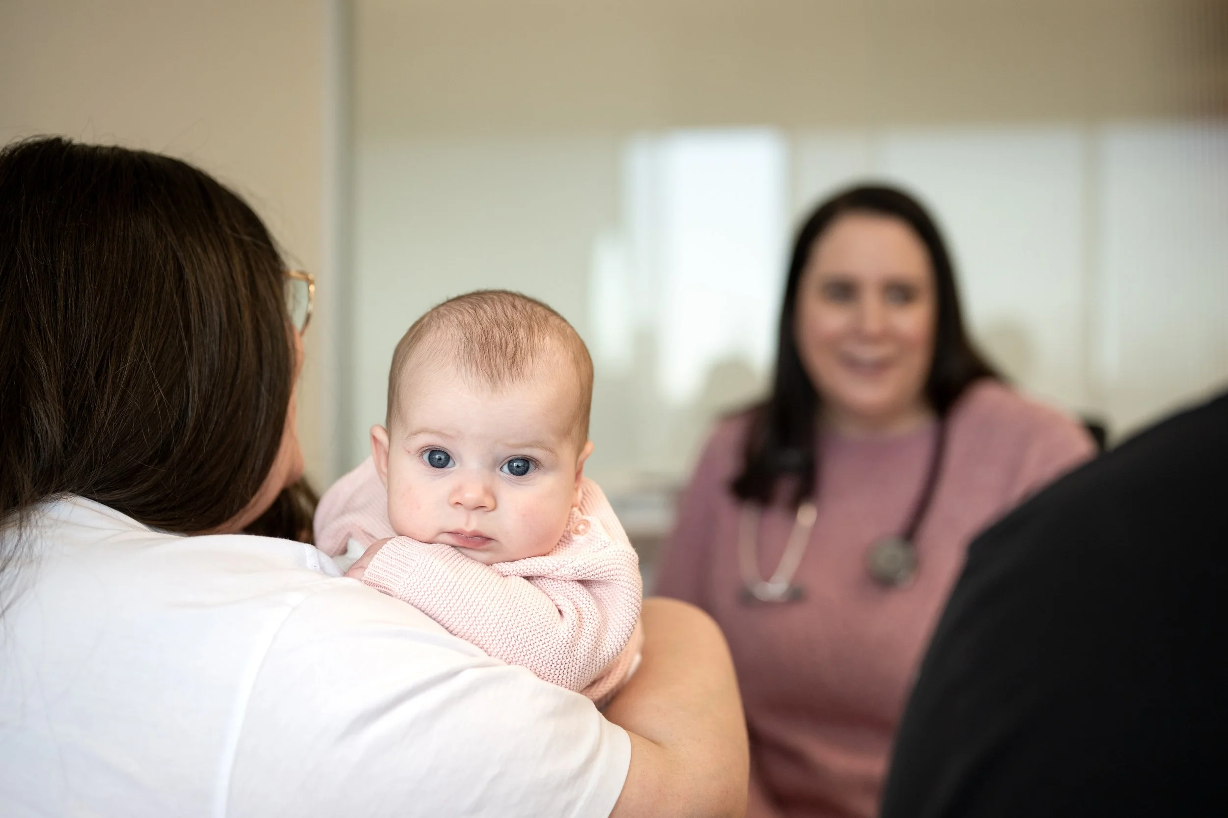 A woman holding a baby with blue eyes, with a woman in the background wearing a stethoscope, in a clinical setting.
