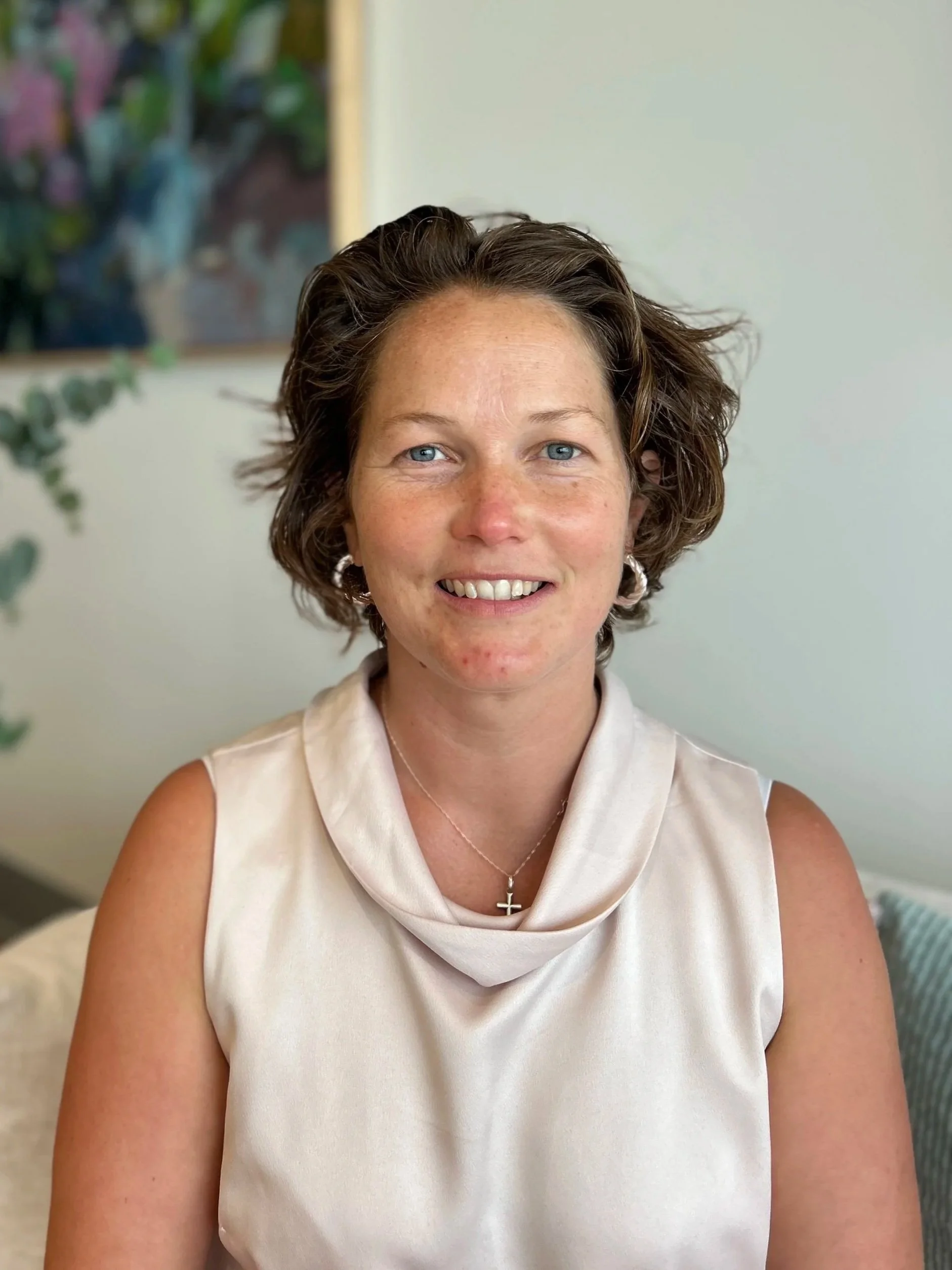 A woman with short, wavy brown hair, blue eyes, and fair skin, smiling, wearing a cream sleeveless top, a silver cross necklace, and hoop earrings, sitting indoors with a blurred background that includes a painting and some plants.