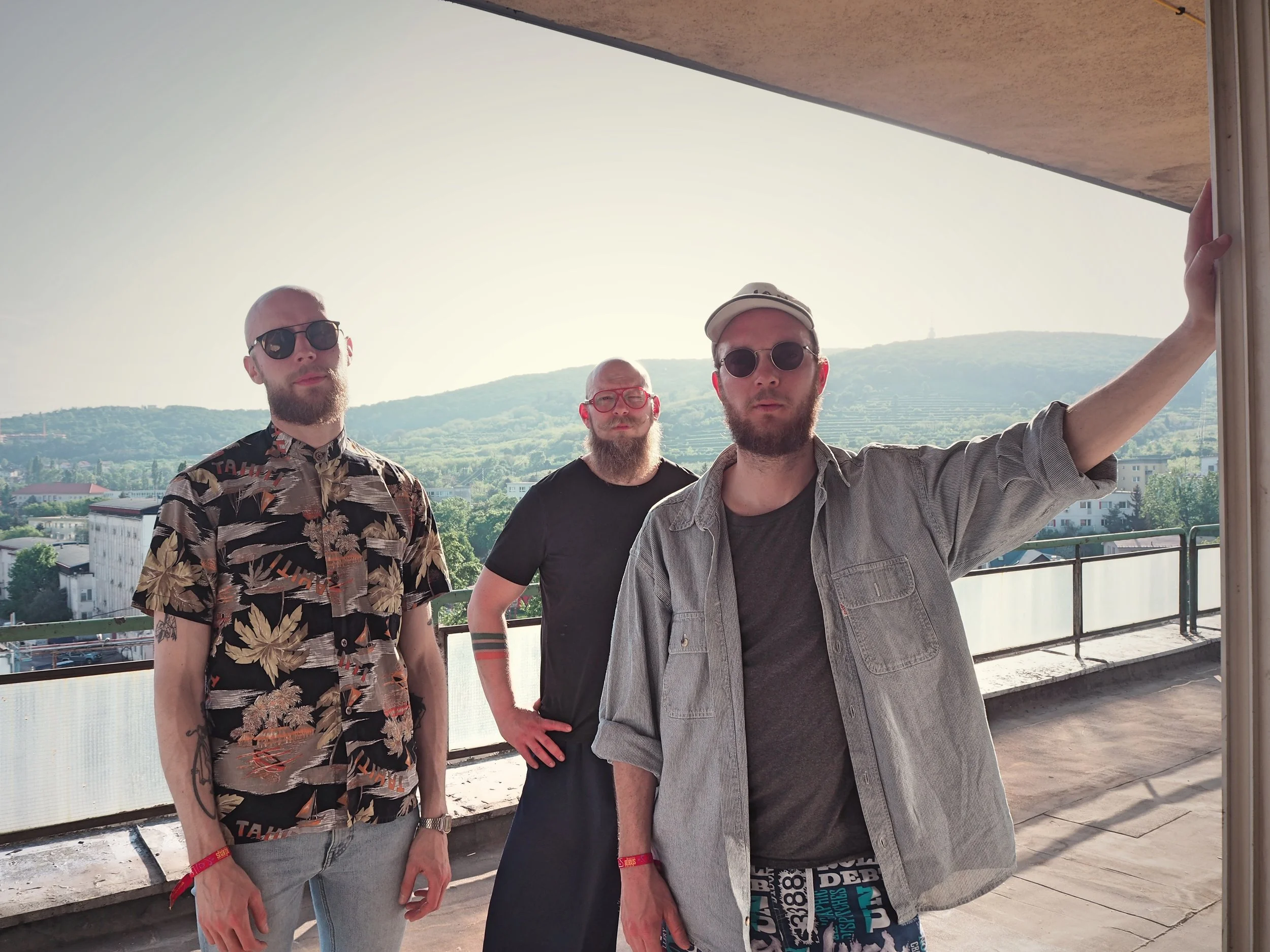 Three men standing on a balcony with a scenic view of green hills, trees, and buildings in the background, all wearing sunglasses on a sunny day.