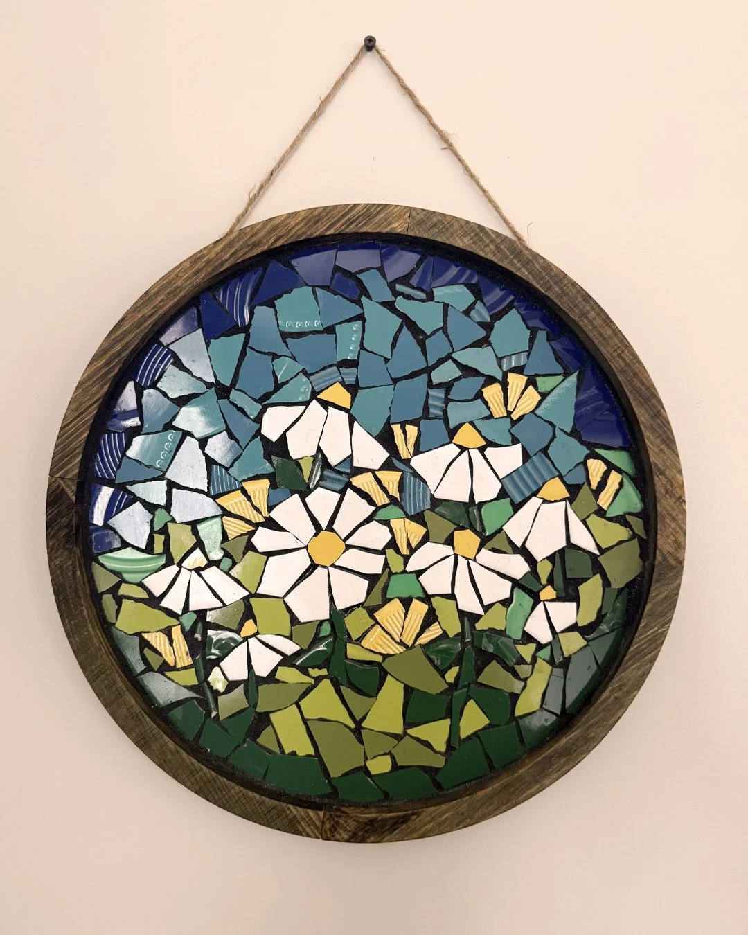Colorful mosaic artwork of white daisies with yellow centers and green leaves in a wooden circular frame hanging on a beige wall.