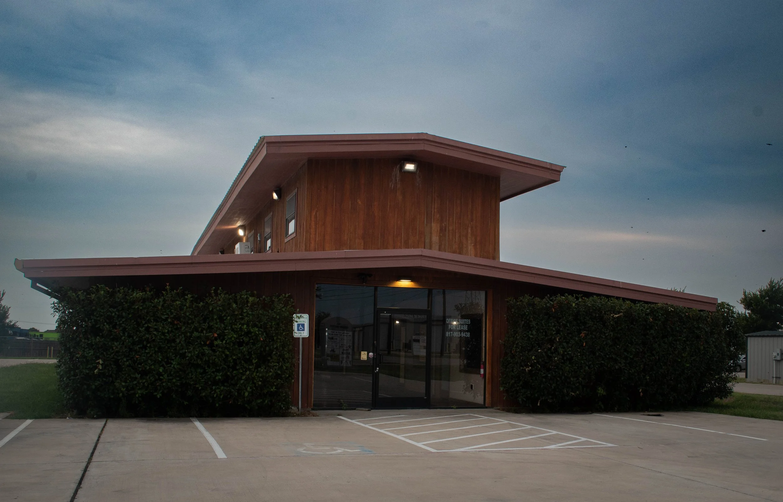 A two-story building with a modern design, featuring wooden exterior walls and a sloped roof. There are outdoor lights on the upper floor, a glass entrance door, and an office for lease sign visible at the entrance. The building is surrounded by neatly trimmed bushes, with an empty parking lot in the foreground, and a cloudy sky overhead.