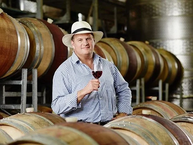 Bruce Jack a winemaker in a checkered shirt and panama hat holding a glass of red wine in a wine cellar with wooden barrels in the background.