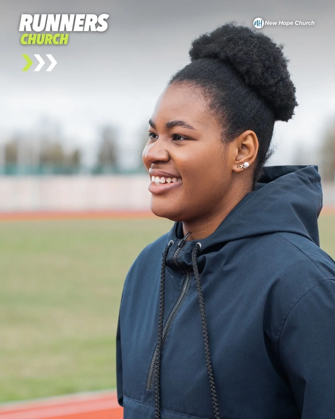 A smiling young Black woman in a navy hooded jacket standing beside an outdoor running track, looking off to her right.
