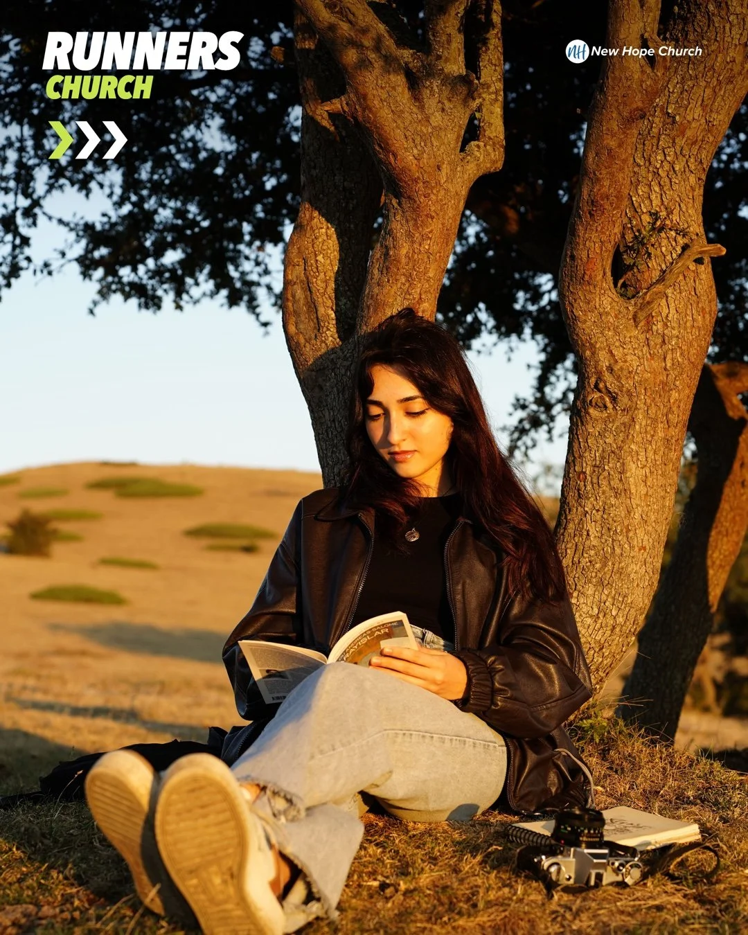 A young woman reads a book beneath a tree in golden evening light, absorbed in what she is reading
