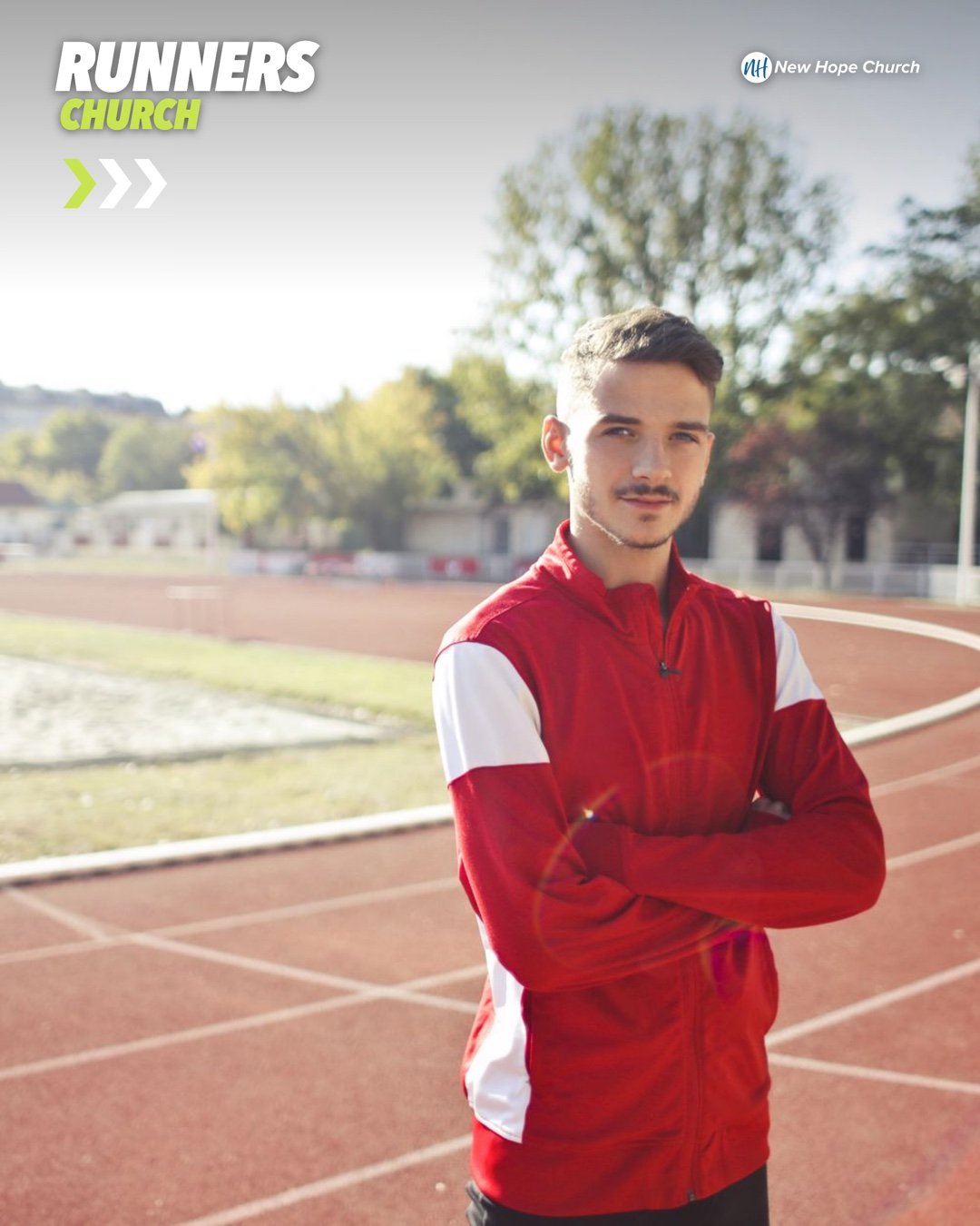 A young male athlete in a red and white track jacket stands with his arms crossed on a sunlit outdoor running track, looking calmly at the camera.