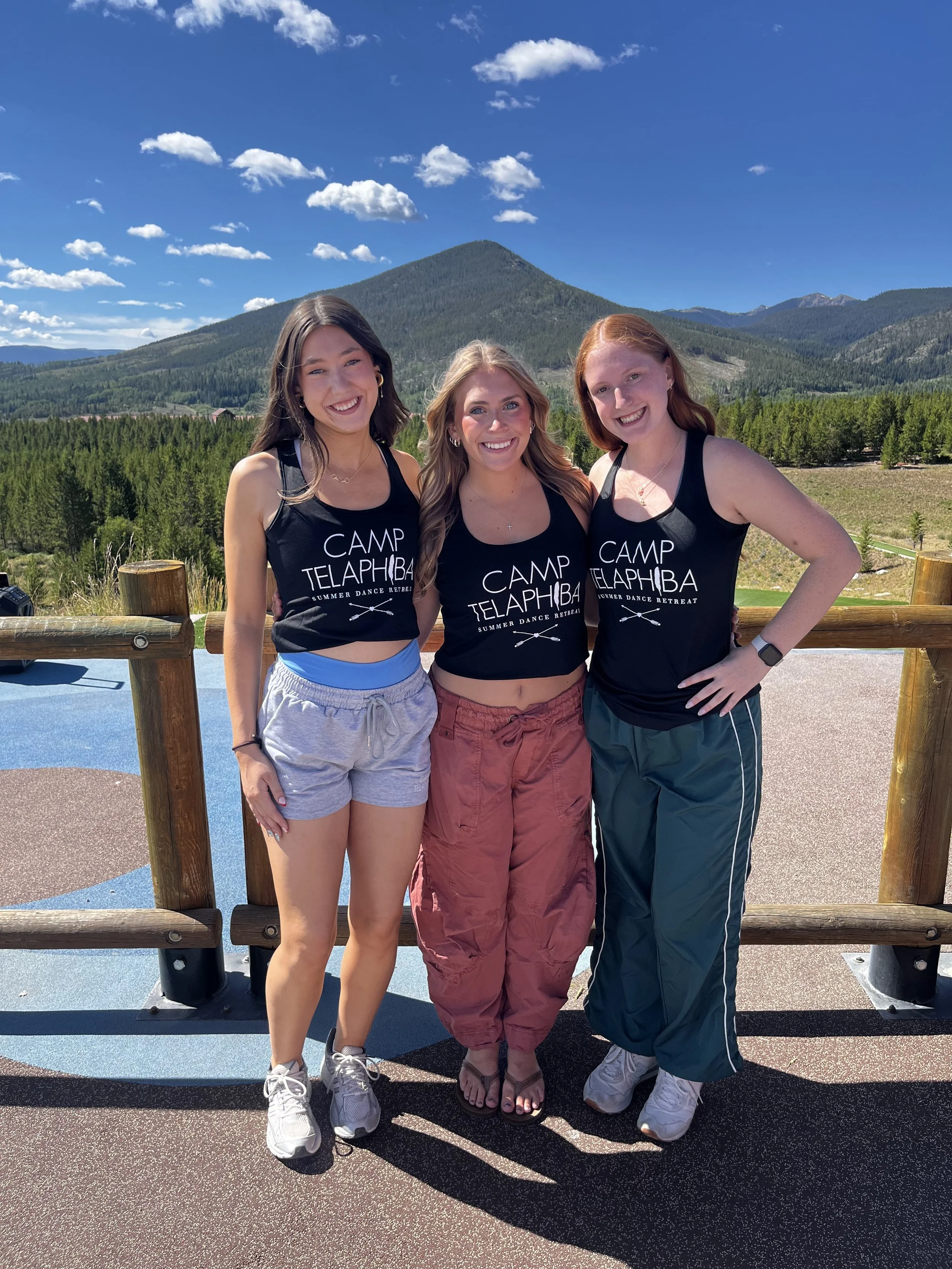 Three young women standing together outdoors, smiling, with a scenic mountainous background and clear blue sky.