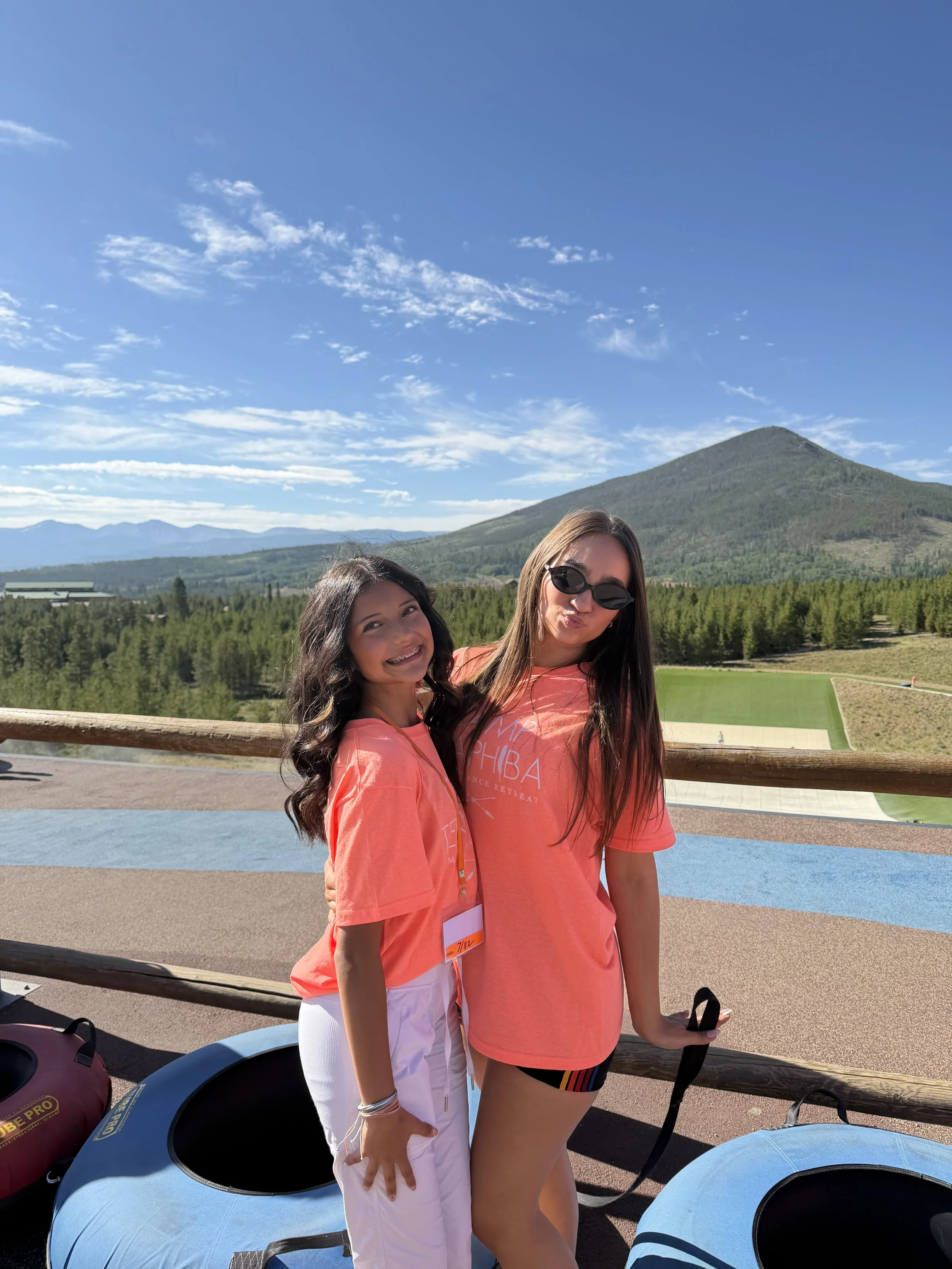 Two women wearing matching orange T-shirts standing outdoors with a scenic mountain landscape, green forest, and blue sky in the background.