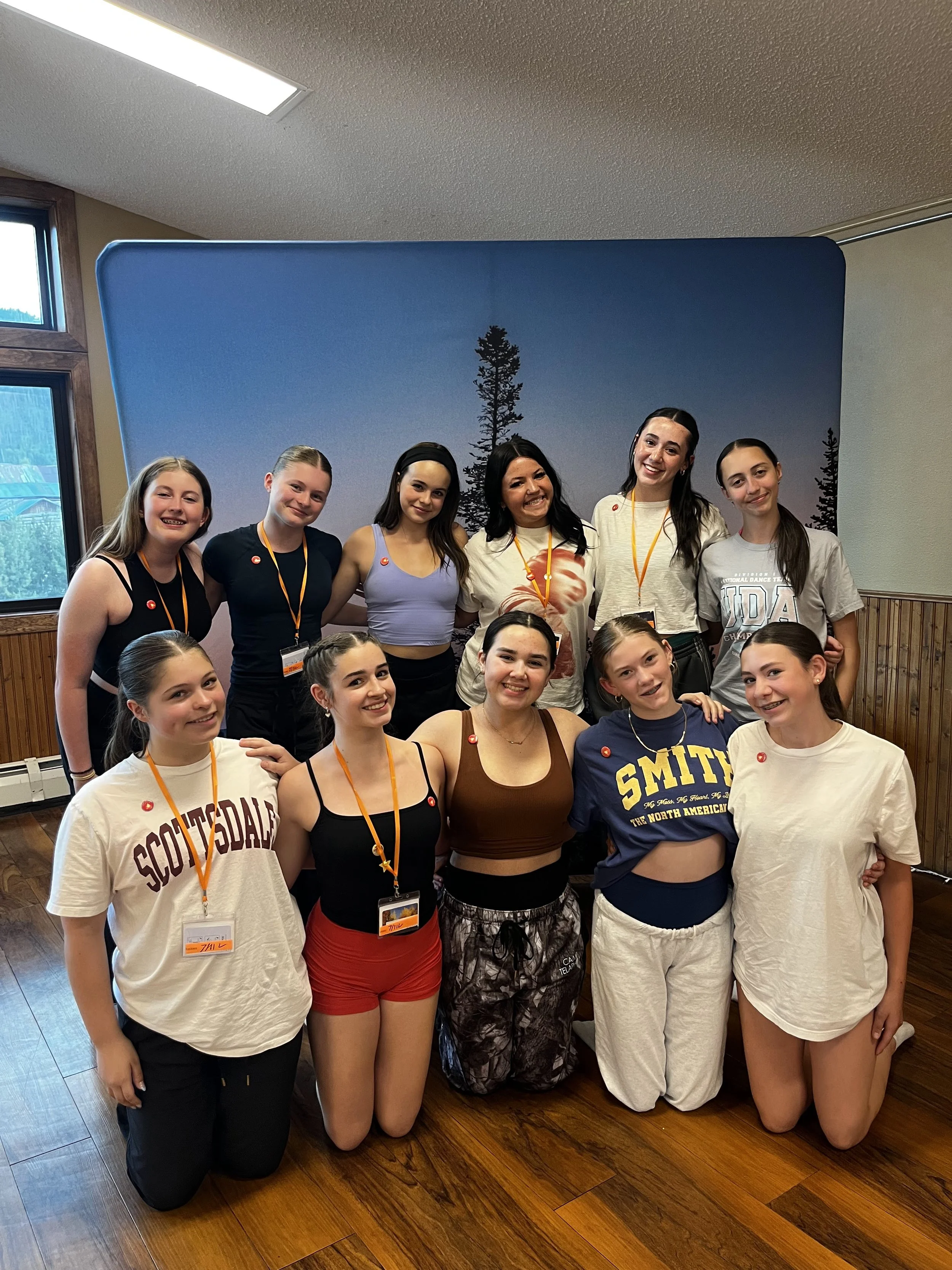 A group of ten young women and girls, some standing and some kneeling, posing together indoors for a photo. They are all wearing casual clothes, with some wearing event badges and lanyards. The background features a blue wall with silhouettes of trees and a window with natural light.