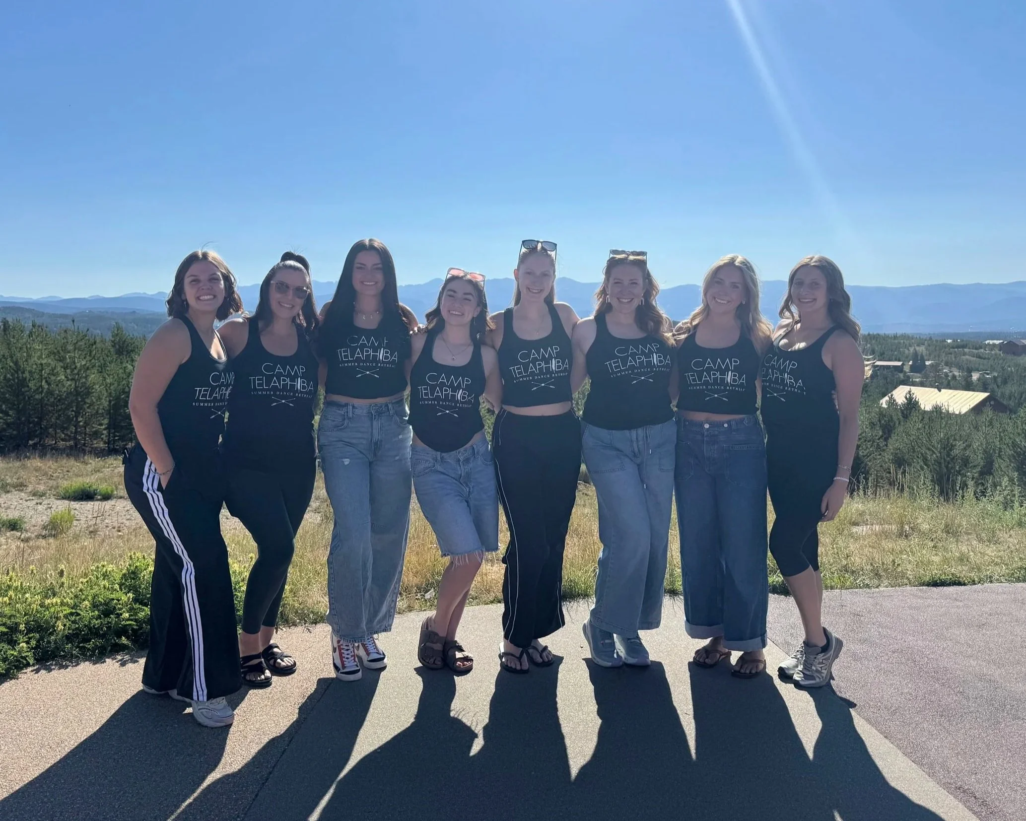 A group of eight young women wearing matching black tank tops that say "CAMP TELAPHI BA" and light blue jeans, standing outdoors on a sunny day with mountains and trees in the background. They are smiling and standing close together, with some wearing sunglasses and sandals.