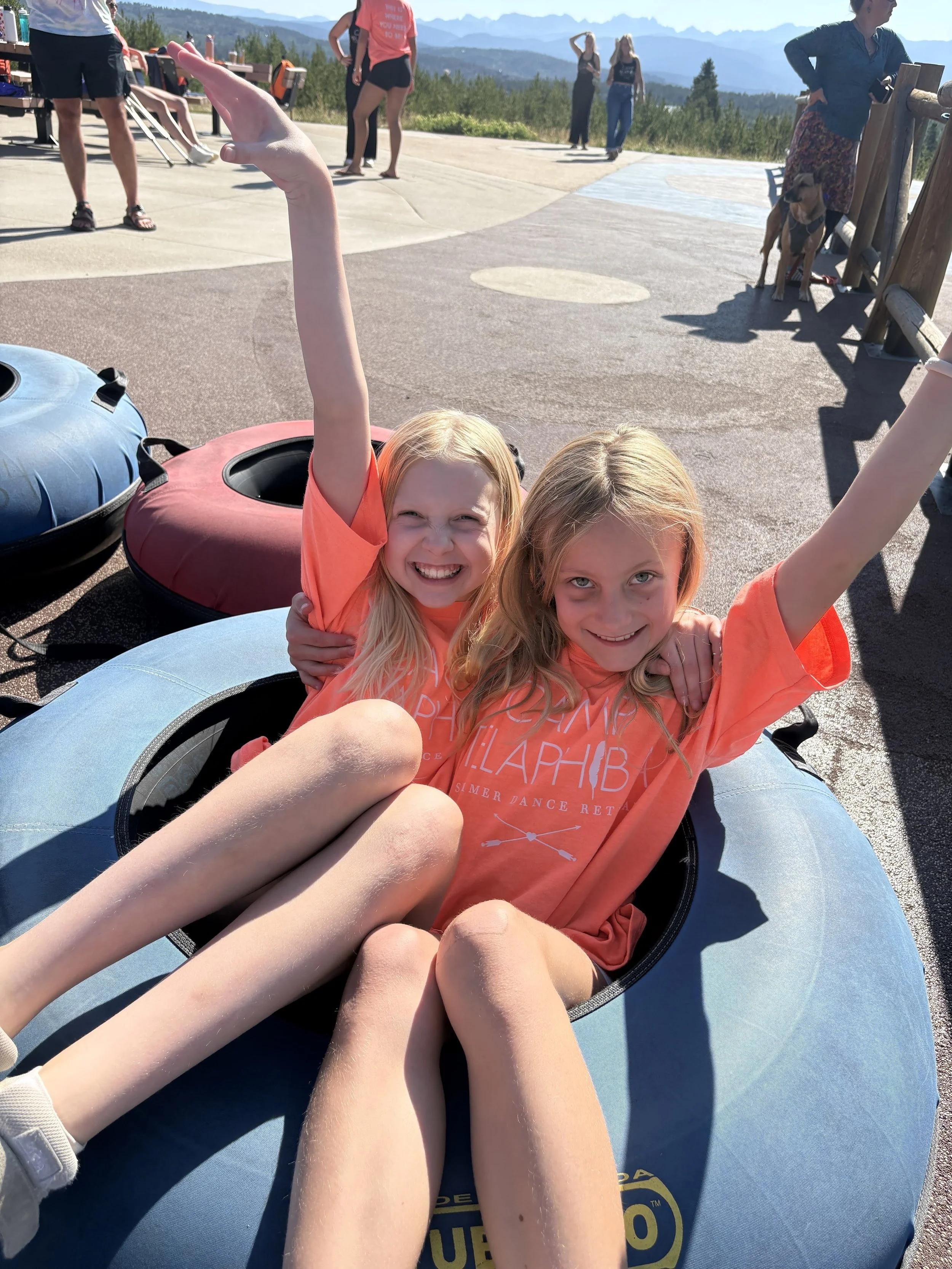 Two young girls sitting in a tire on an outdoor viewing platform, smiling and waving at the camera with arms raised. Several people are standing or walking in the background with scenic mountains and trees visible under a clear sky.