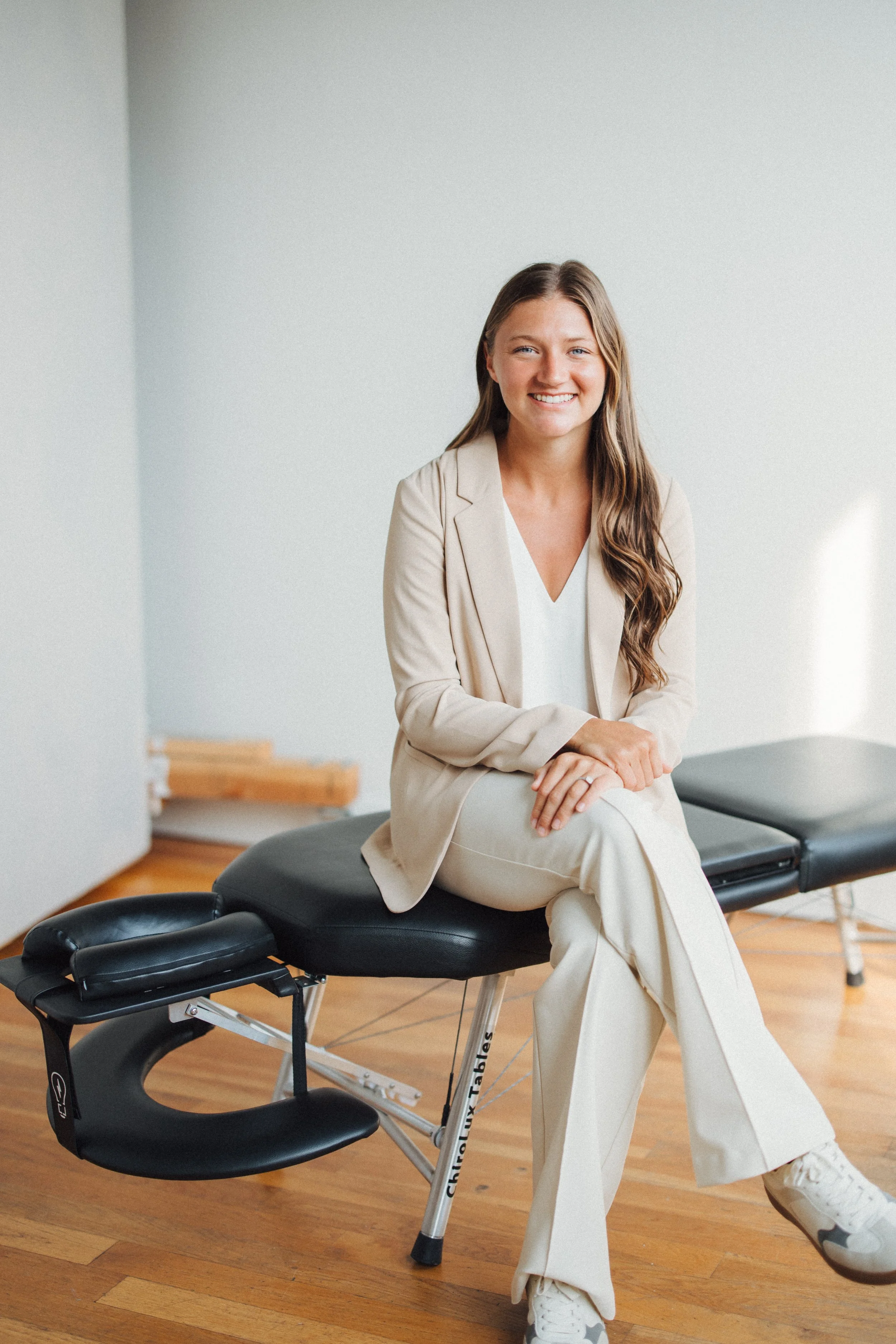 A woman with long brown hair, wearing a beige blazer and white outfit, sitting on a chiropractic table in a medical office, smiling at the camera.