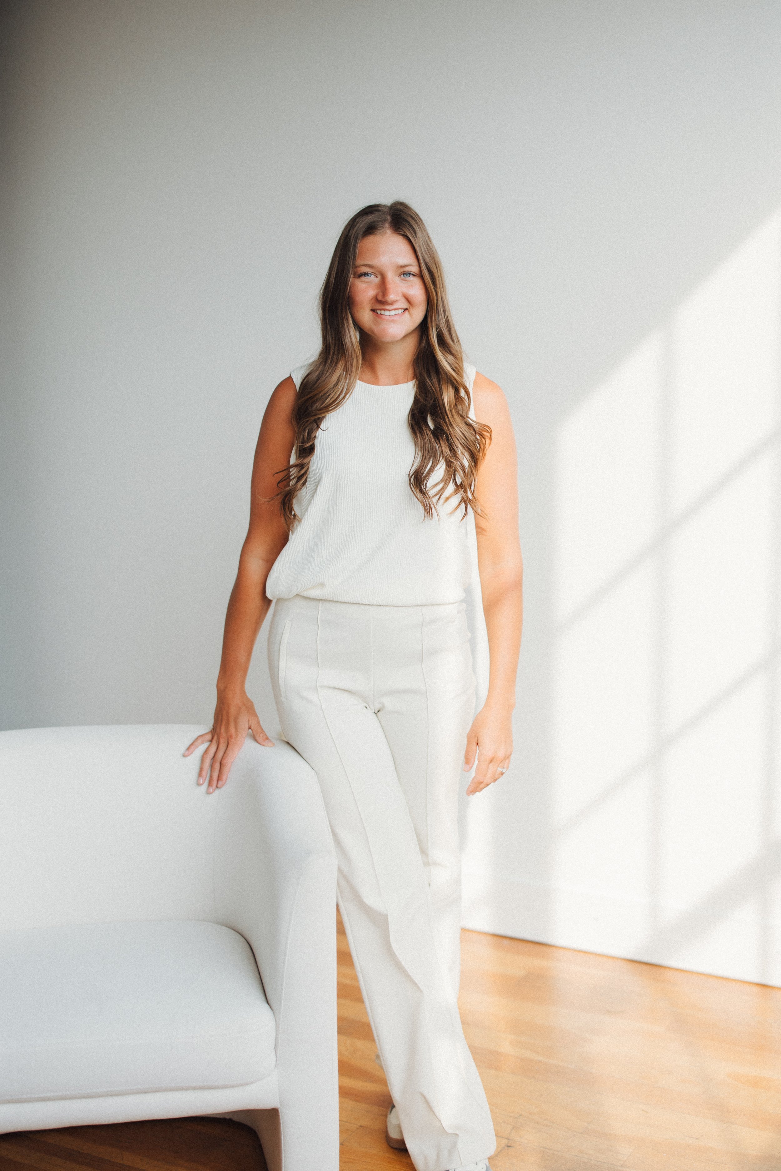 Woman with long brown hair smiling, standing next to a white sofa in a bright room with white walls and wooden floor.