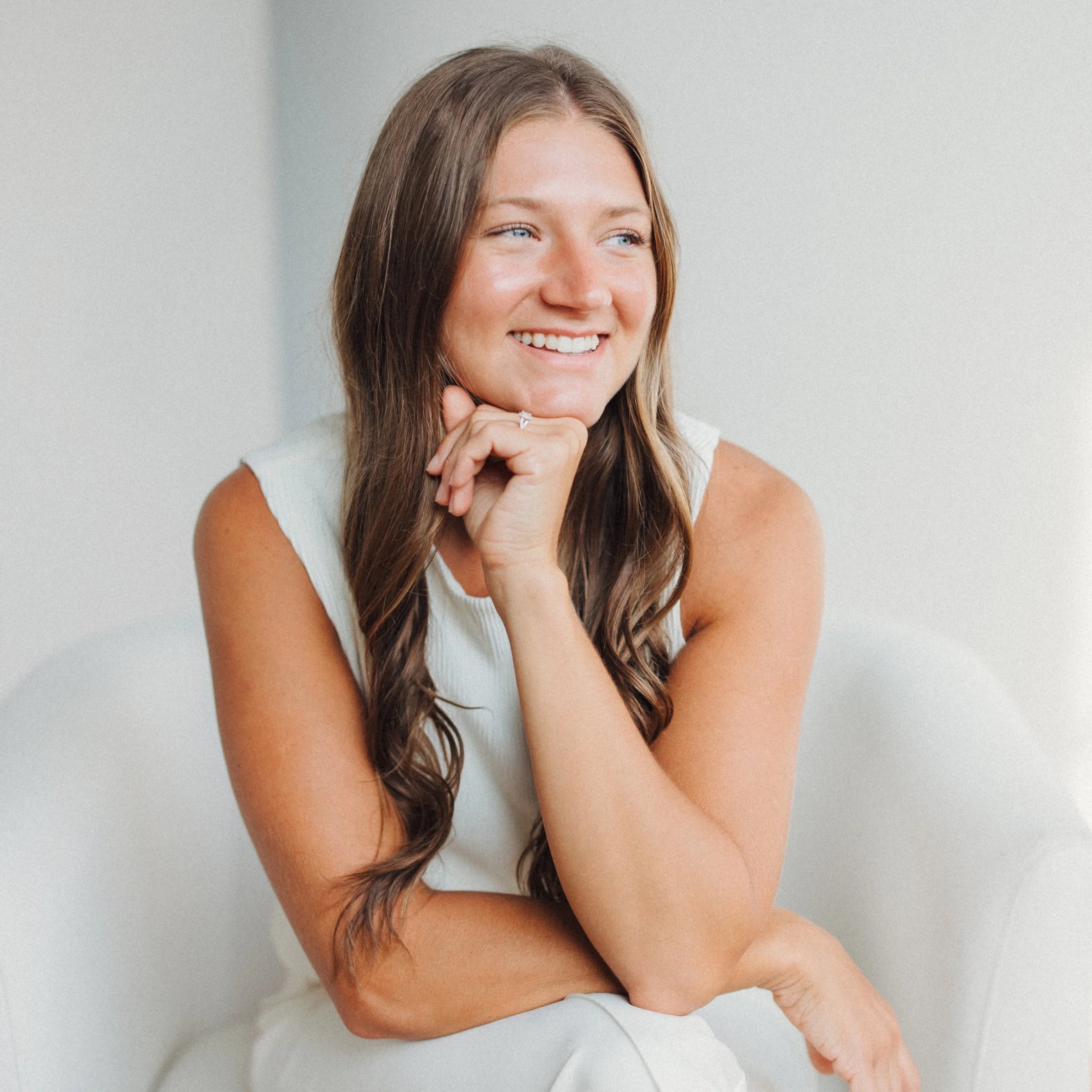 A smiling young woman with long brown hair, wearing a white sleeveless top, sitting on a white chair, resting her chin on her hand.