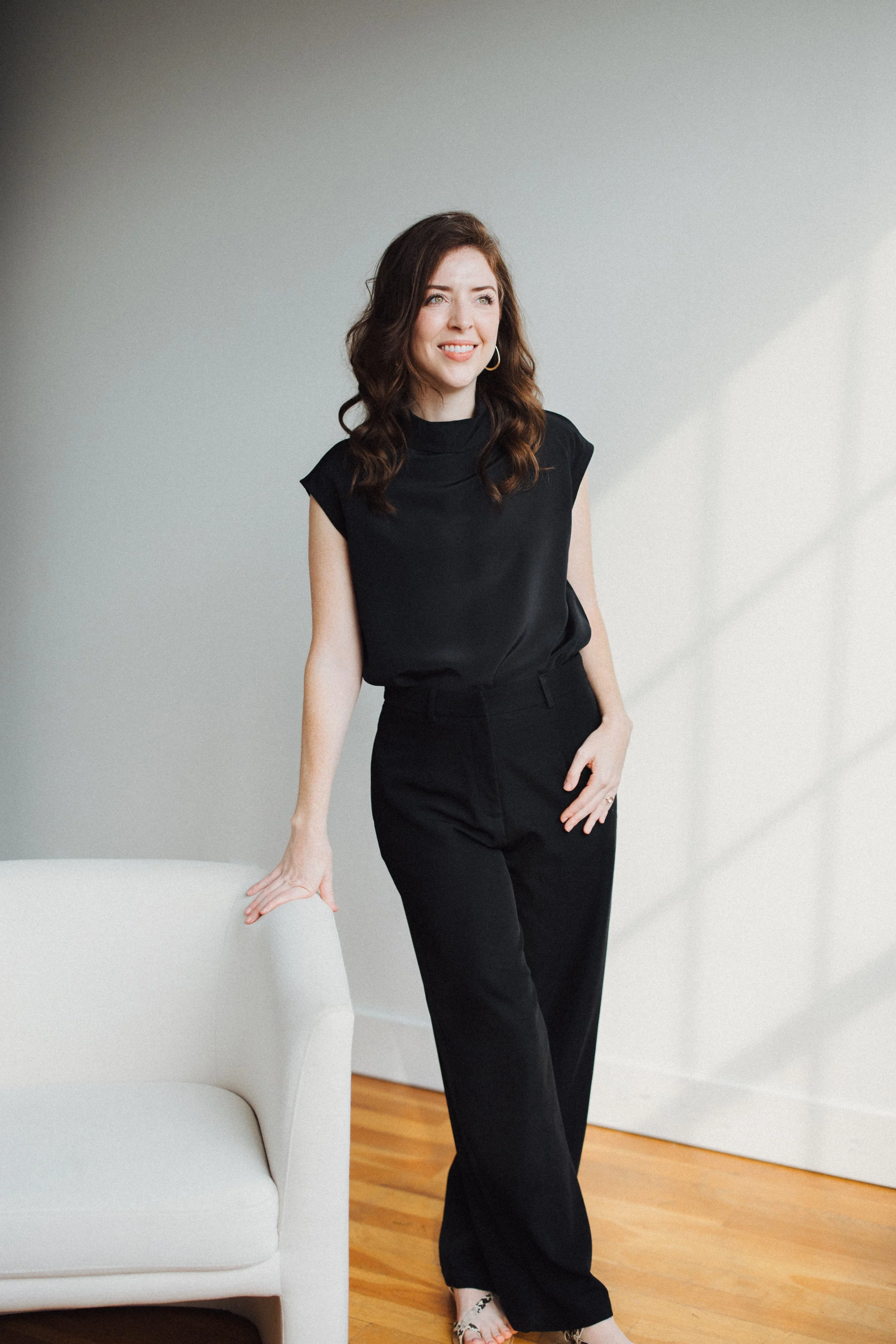 A woman in black attire standing near a white sofa in a room with wooden flooring and white walls, smiling.