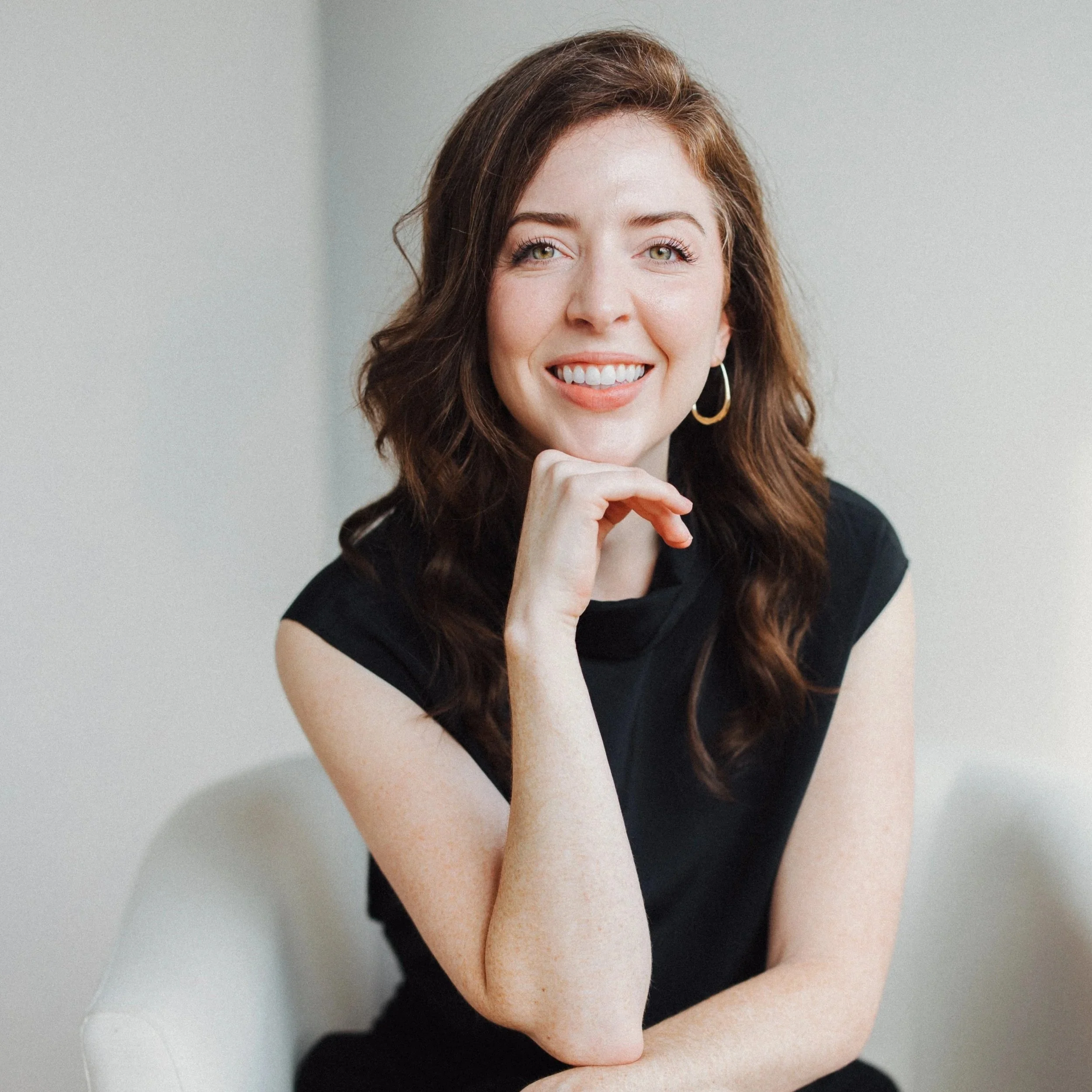 A woman with wavy brown hair, wearing a black sleeveless top and gold hoop earrings, smiling and resting her chin on her hand against a neutral background.