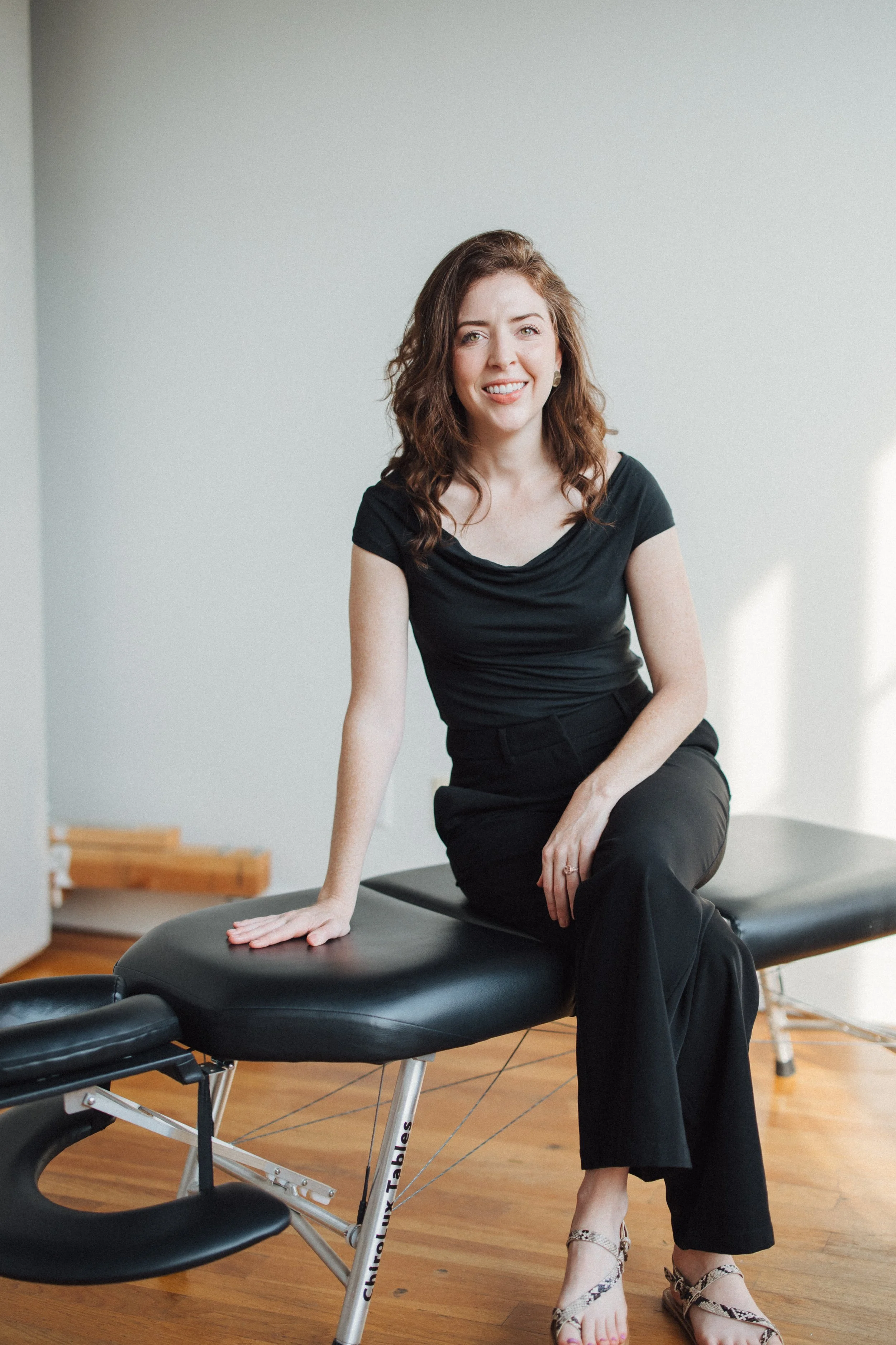 A woman with wavy brown hair, wearing a black top and black pants, sitting on a chiropractic table in a room with wooden flooring and a plain white wall, smiling at the camera.