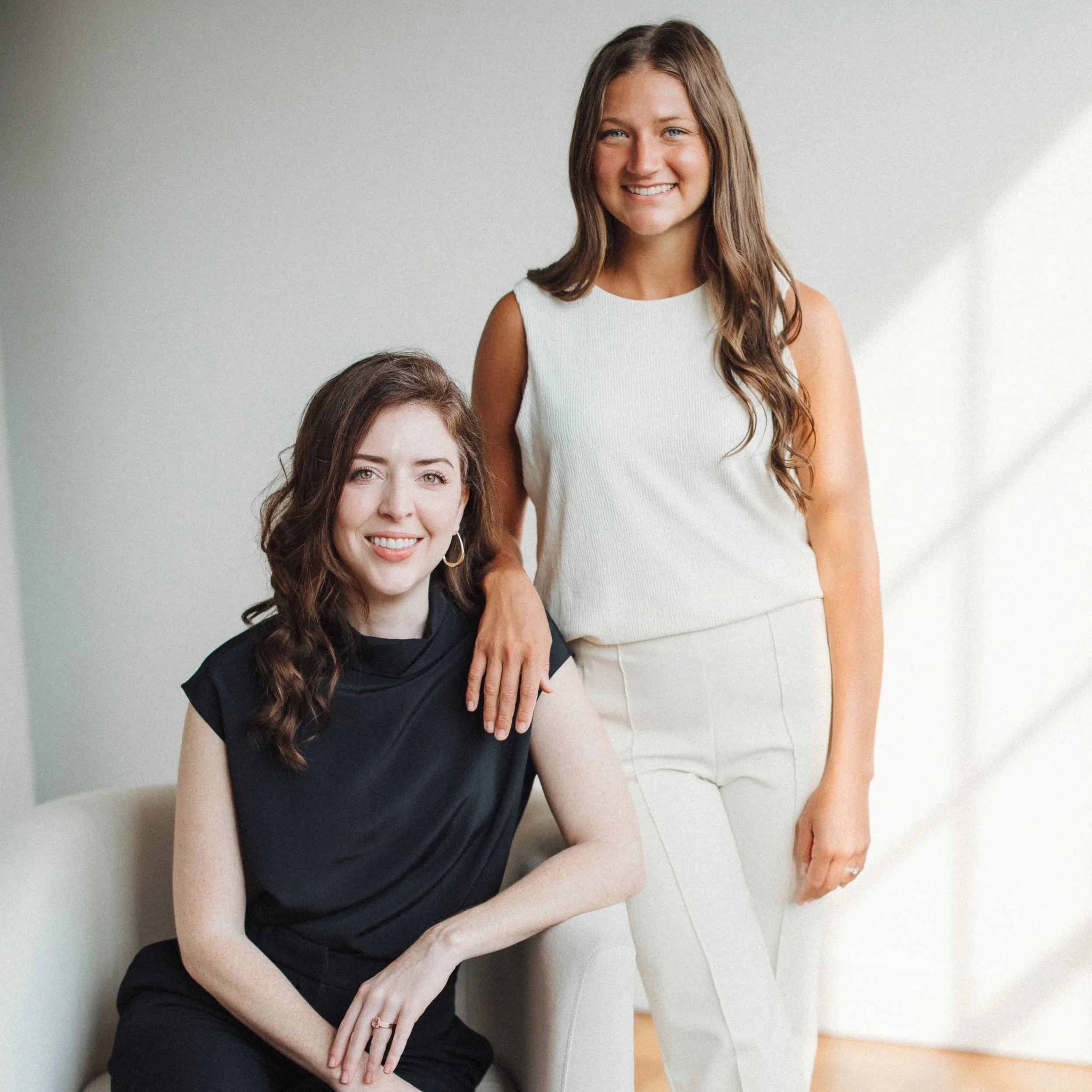 Two women with brown hair and fair skin, one seated and one standing, smiling in a well-lit room with a white wall background.
