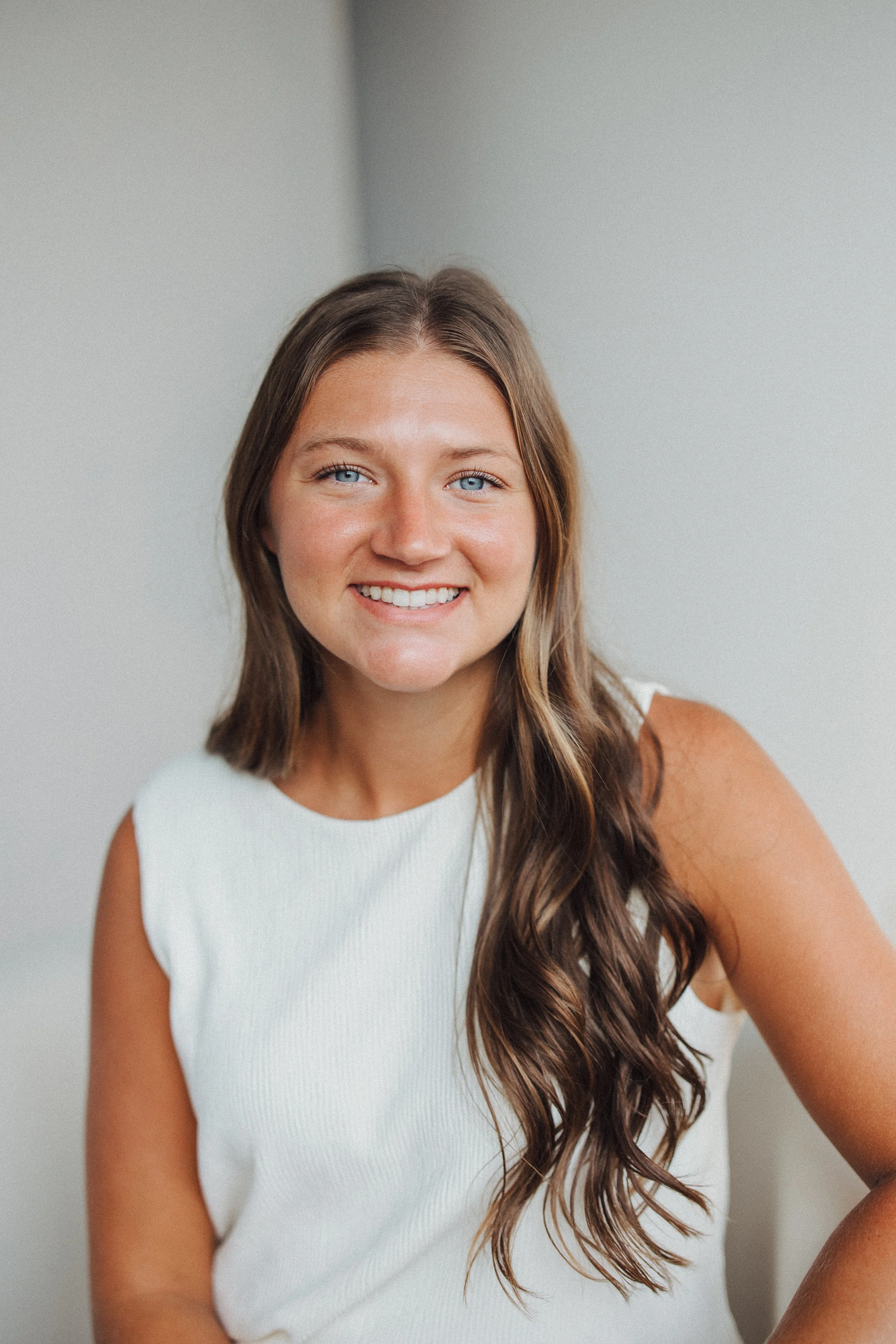 A smiling woman with long, wavy brown hair wearing a sleeveless white top, sitting against a light gray wall.