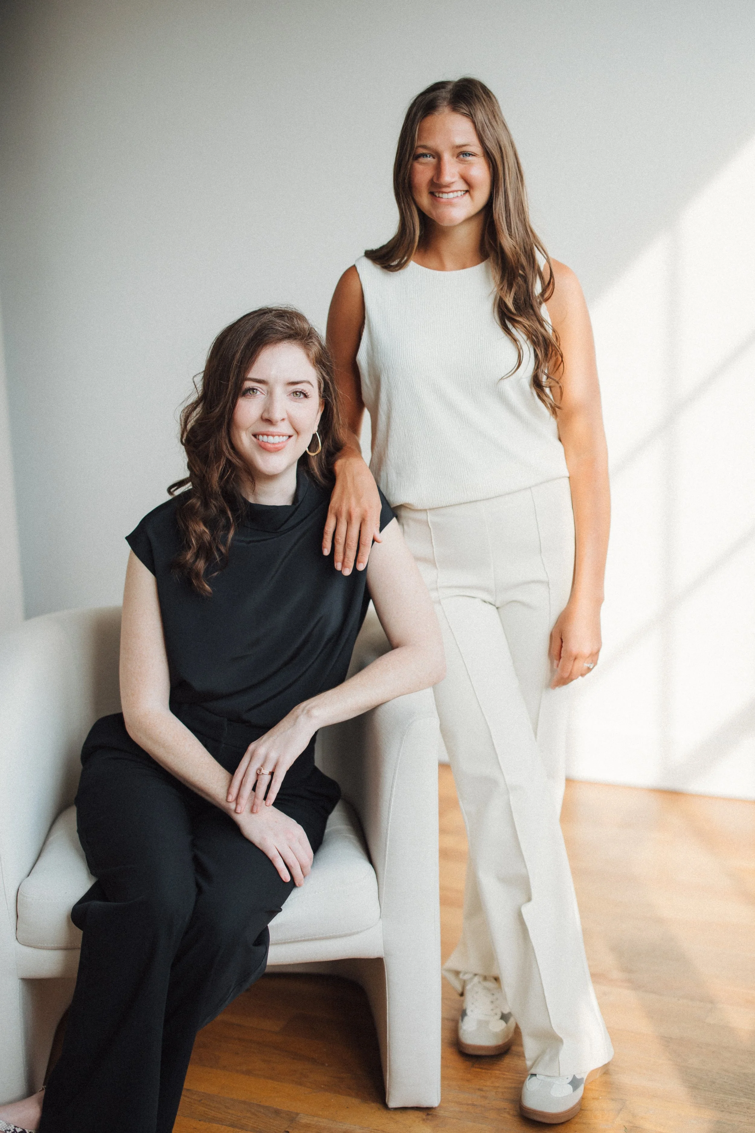 Two women in neutral-colored clothing posing in a minimalist room with wooden floors, one seated on a chair and the other standing beside her, both smiling.