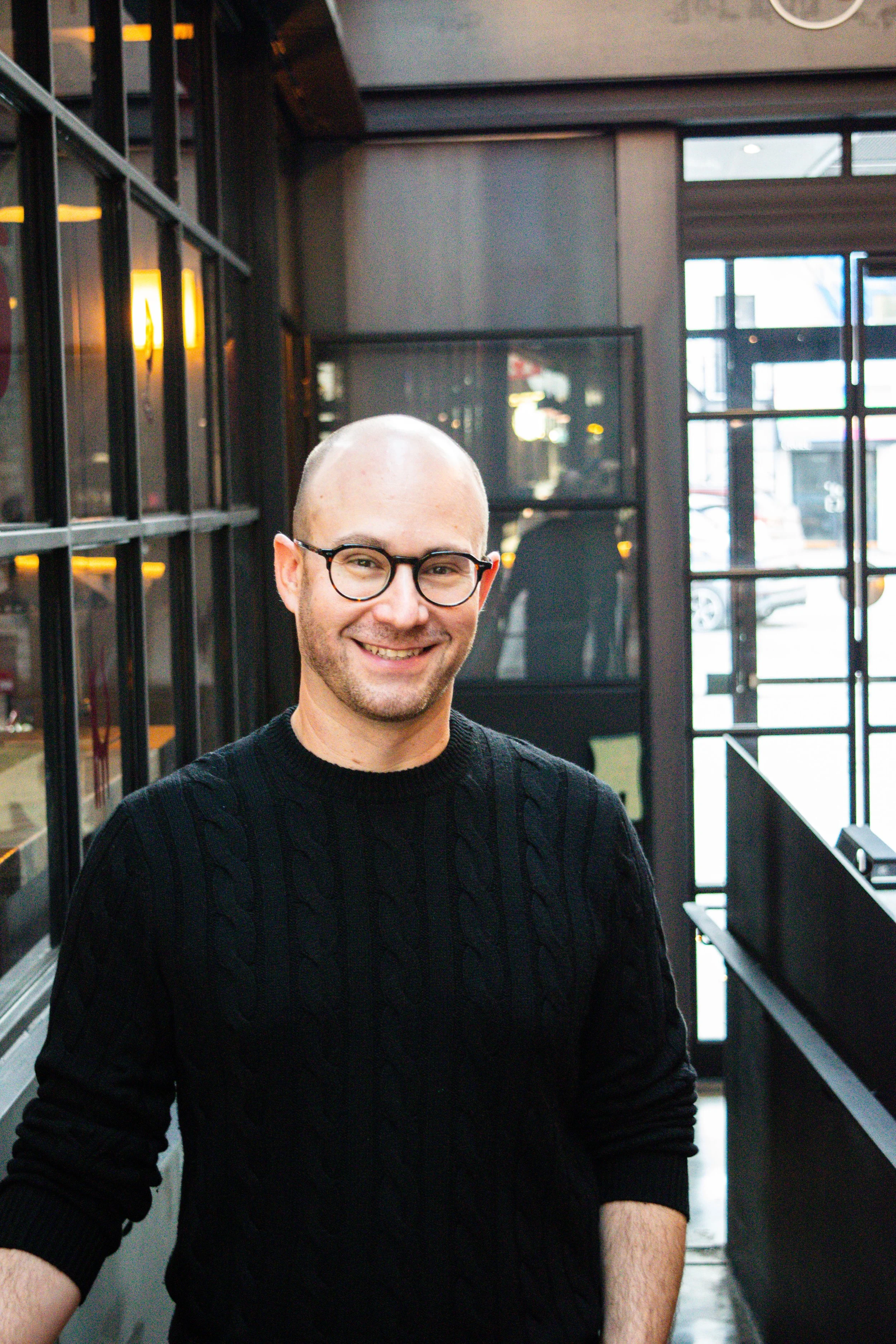 Smiling person in a black sweater standing in a modern interior with glass windows.