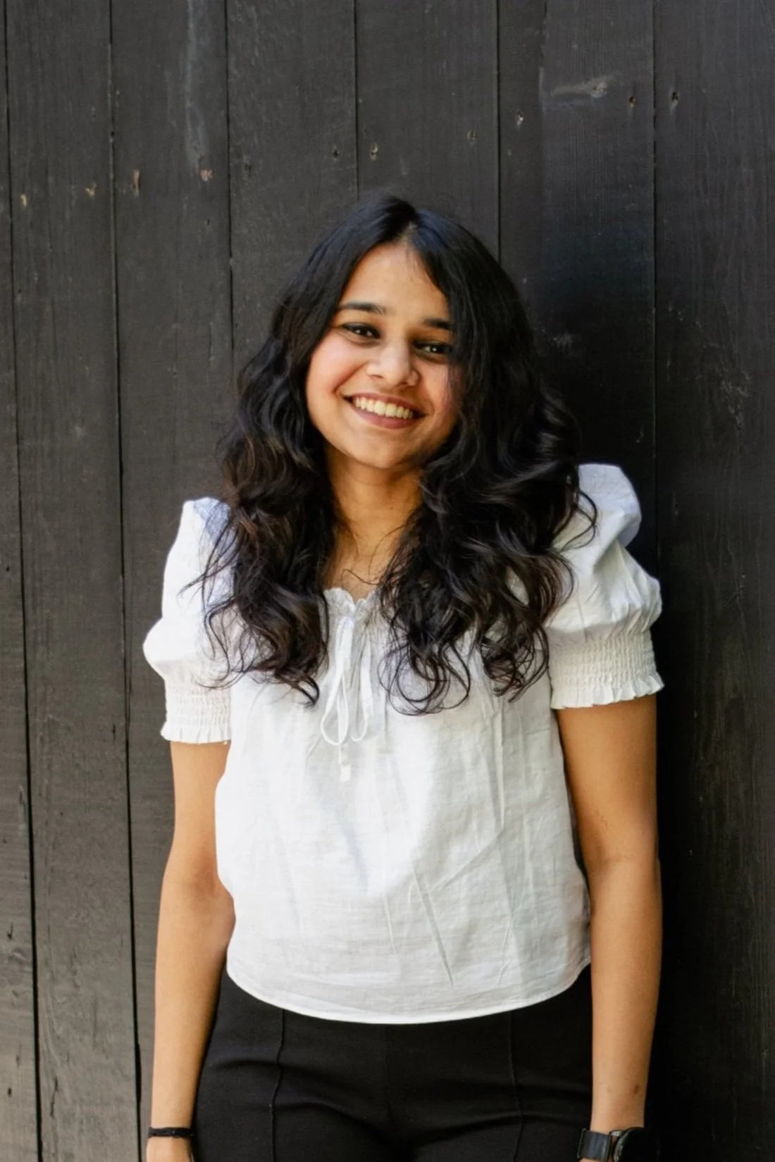 Smiling woman with long curly hair in a white blouse standing against a dark wooden wall.