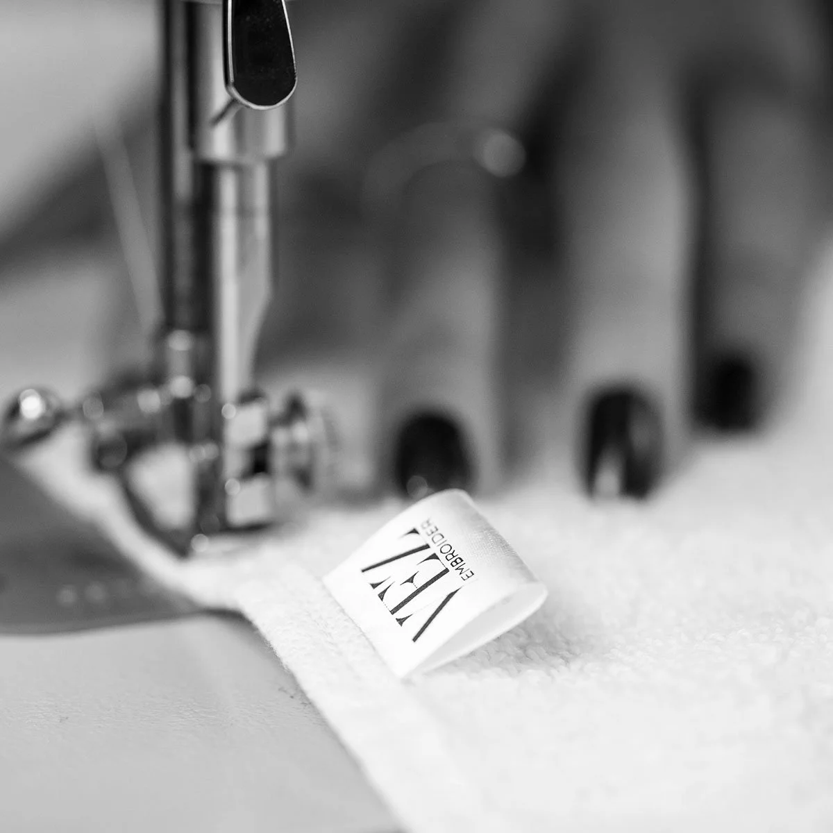 Close-up of a sewing machine stitching a fabric label with the text "VEZEMBROIDER" on white fabric.