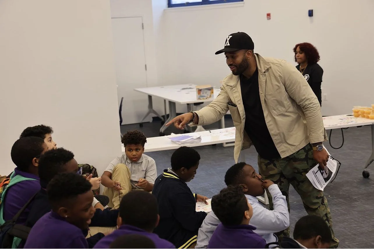 Man engaging with a group of children seated on the floor in a classroom or community space, with a woman in the background. The man is pointing and holding papers, while the children listen attentively.
