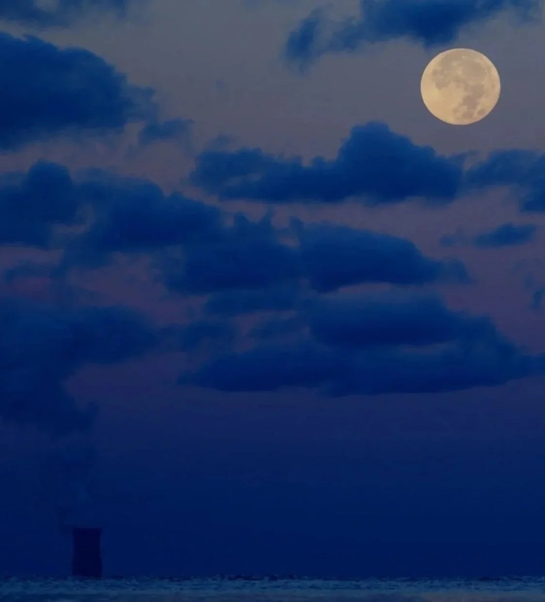 Full moon in a cloudy night sky over a smokestack emitting smoke or steam.
