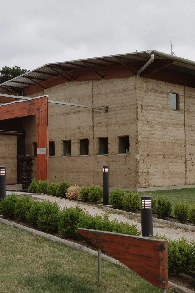 Modern building with concrete walls, small windows, a metal roof, and a landscaped walkway with bushes and black bollard lights.