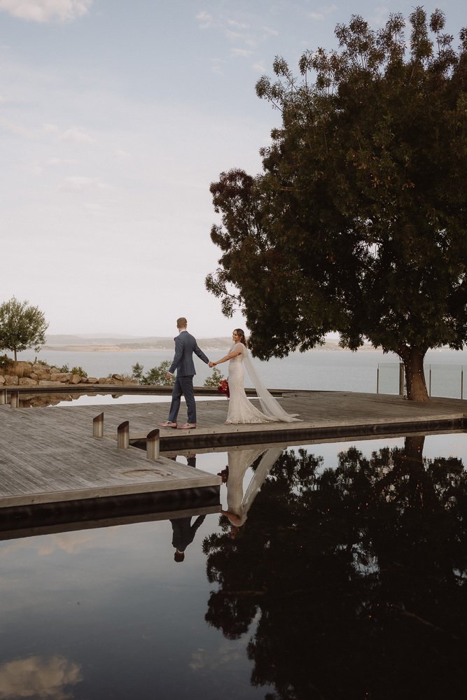 A bride and groom holding hands standing on a wooden dock near a body of water, with a large tree and a cloudy sky in the background.