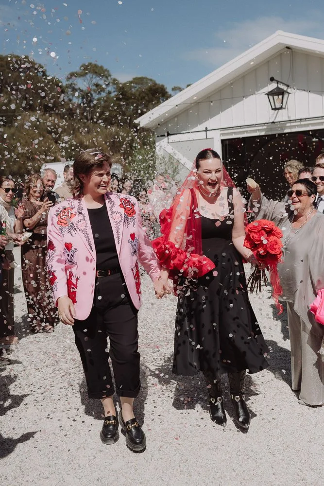Celebrating a wedding with two women walking hand-in-hand through a confetti shower, surrounded by friends and family outside a white building under a clear blue sky.