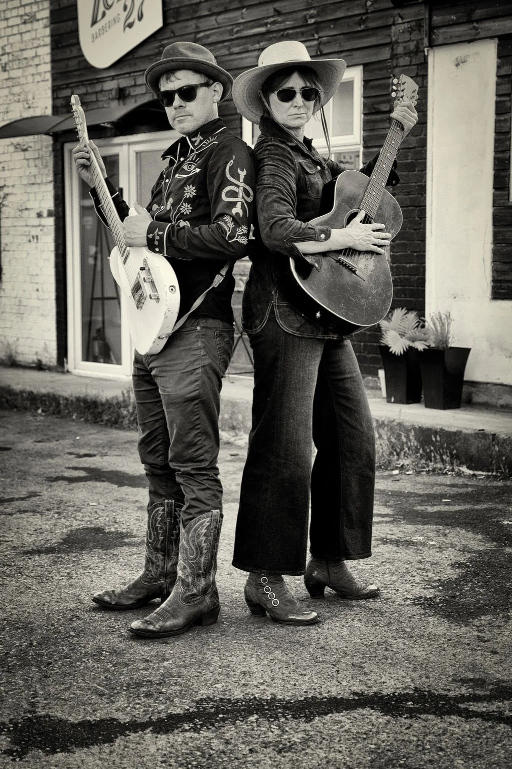 Two musicians standing back-to-back outdoors, both playing guitars. They wear sunglasses, cowboy hats, and western-style clothing, with a brick building in the background.