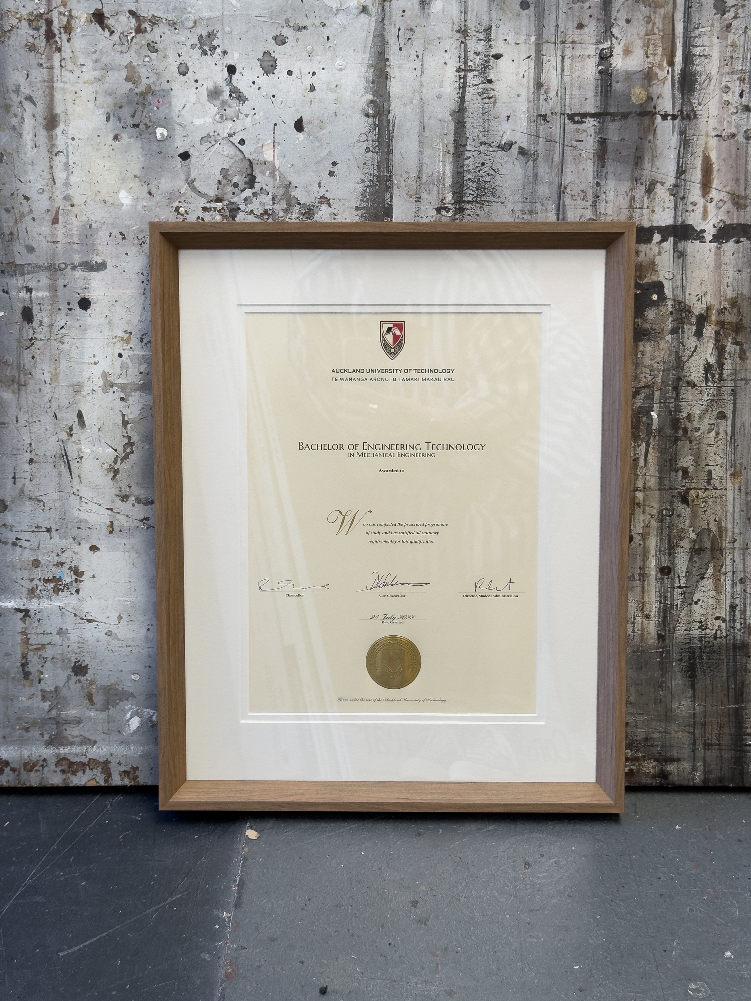 Framed degree from Auckland University of Technology awarded to Bachelor of Engineering Technology in Mechanical Engineering. It has a mid toned wood frame in an angled shape around it, and an off white mat. 