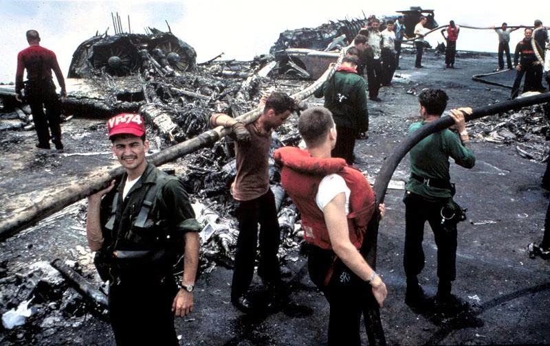Grimy and exhausted, crewmen of USS Forrestal and her airwing continue firefighting efforts in the aftermath of the blaze that killed 134 of their shipmates 29 July 1967. The ship was on station off the coast of Vietnam. In the background are what remains of a row of F-4B Phantoms that were parked along the starboard stern quarter. (Navy released image.)