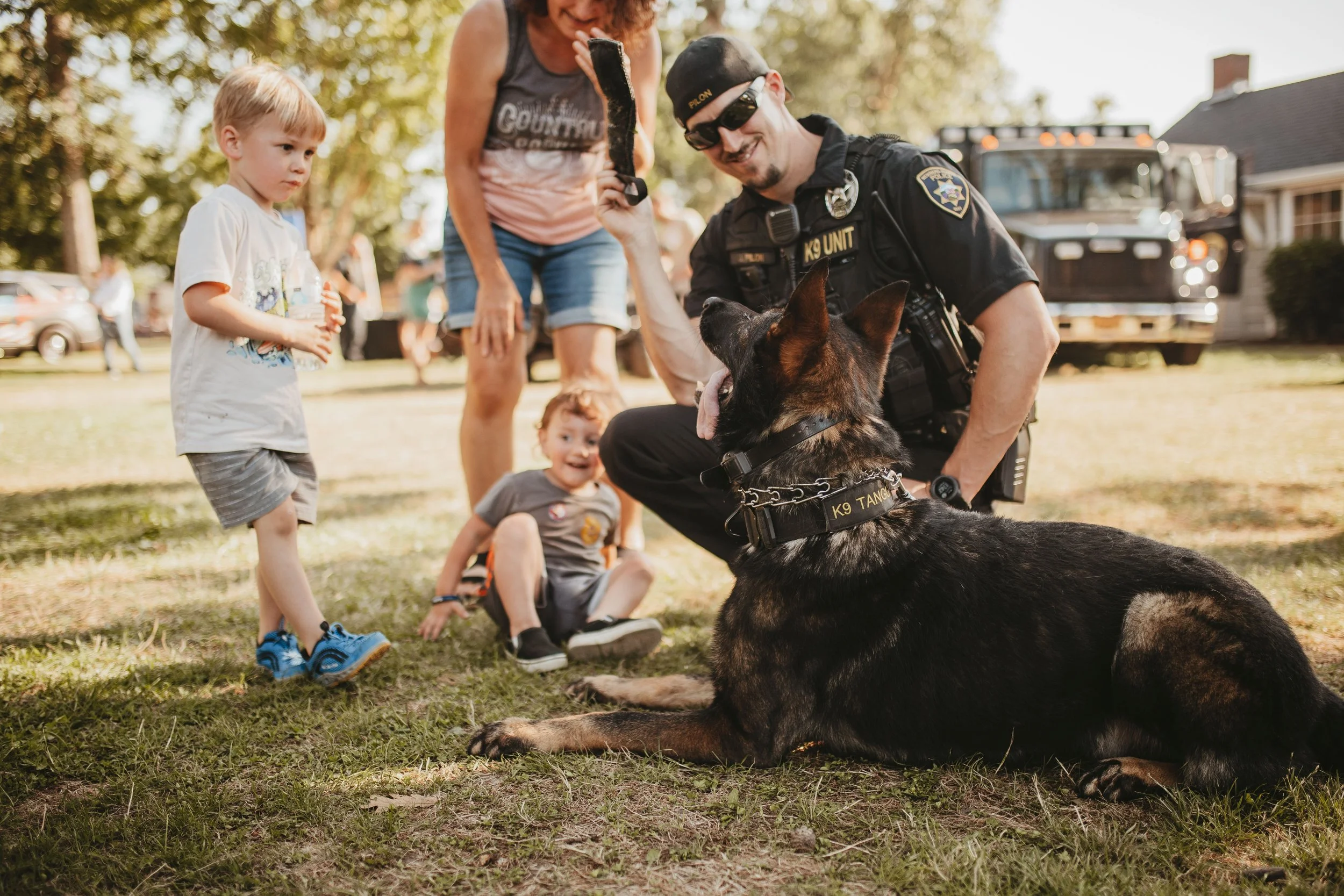 A police officer kneeling with a police dog, greeting two young children in a park, with people and a truck in the background on a sunny day.