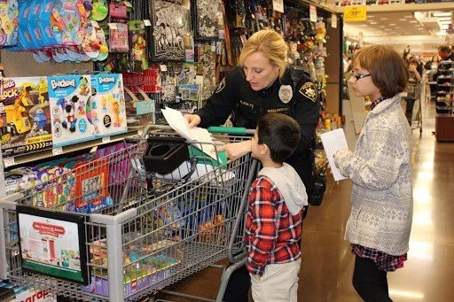 A police officer is talking to two children at a store aisle, with a shopping cart in front of them filled with toys and supplies. The officer appears to be showing or explaining something on a piece of paper.