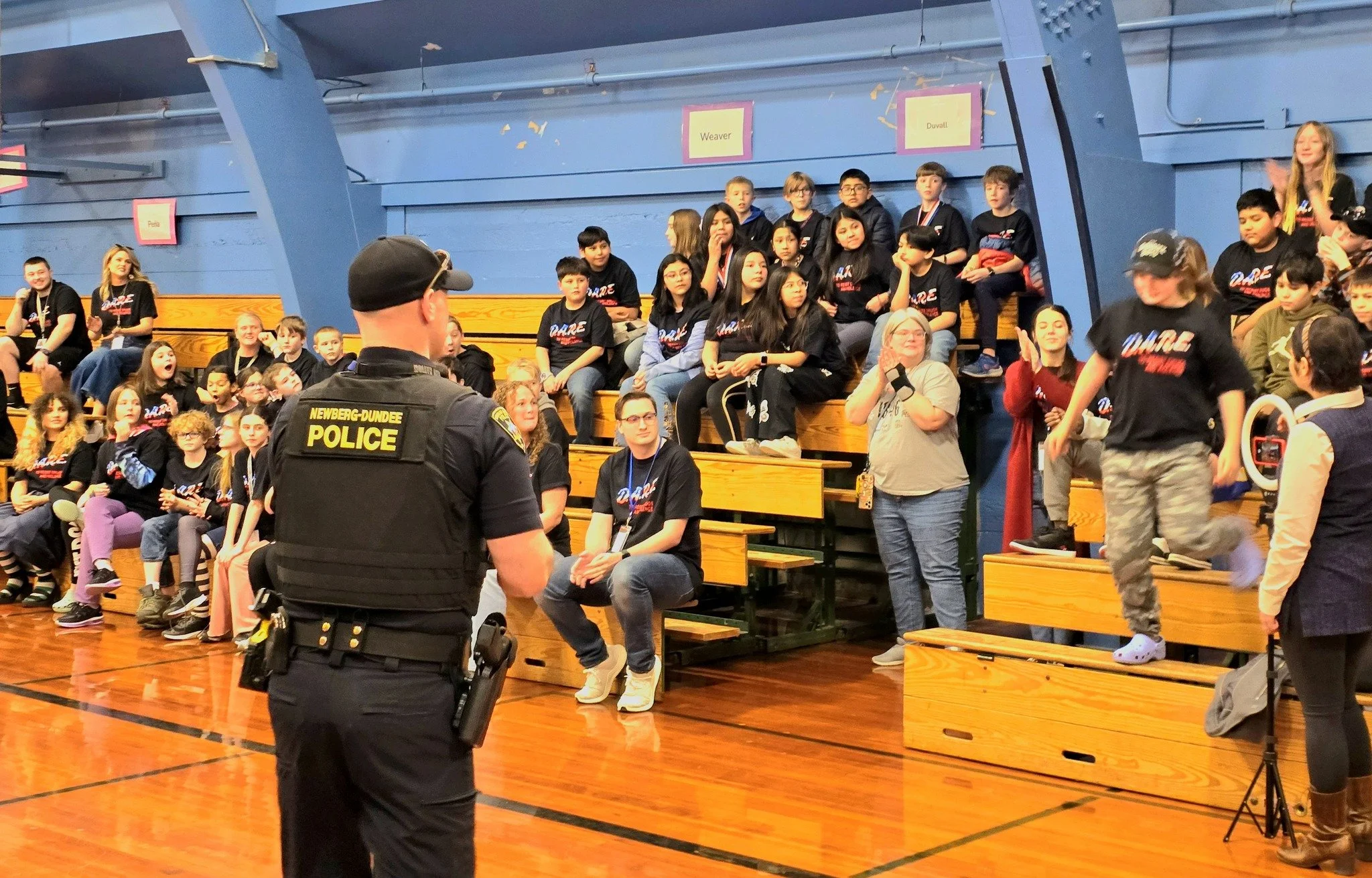 A police officer from Newberg-Dundee Police stands in front of a large group of children and adults in a gymnasium. The audience is seated on wooden bleachers, and some children are standing on the gym floor.