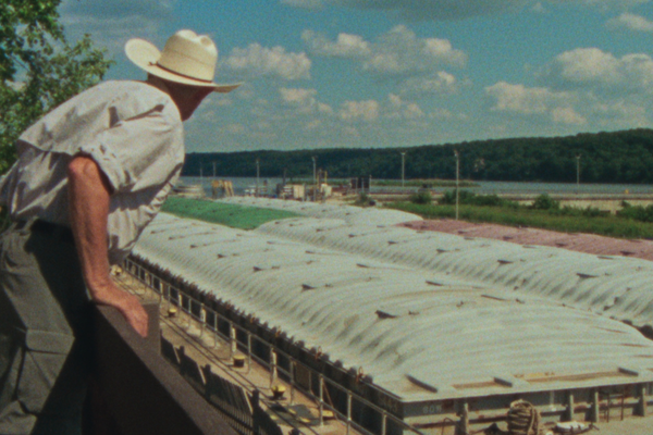 Still of man in hat looking at barges on a river from AMERICAN DENDRITE.