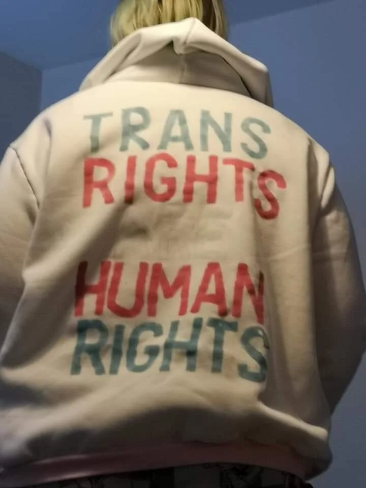 The back of white hooded sweatshirt worn by a blonde haired person. The words “Trans rights are human rights” have been painted on the back in pink, blue and white.