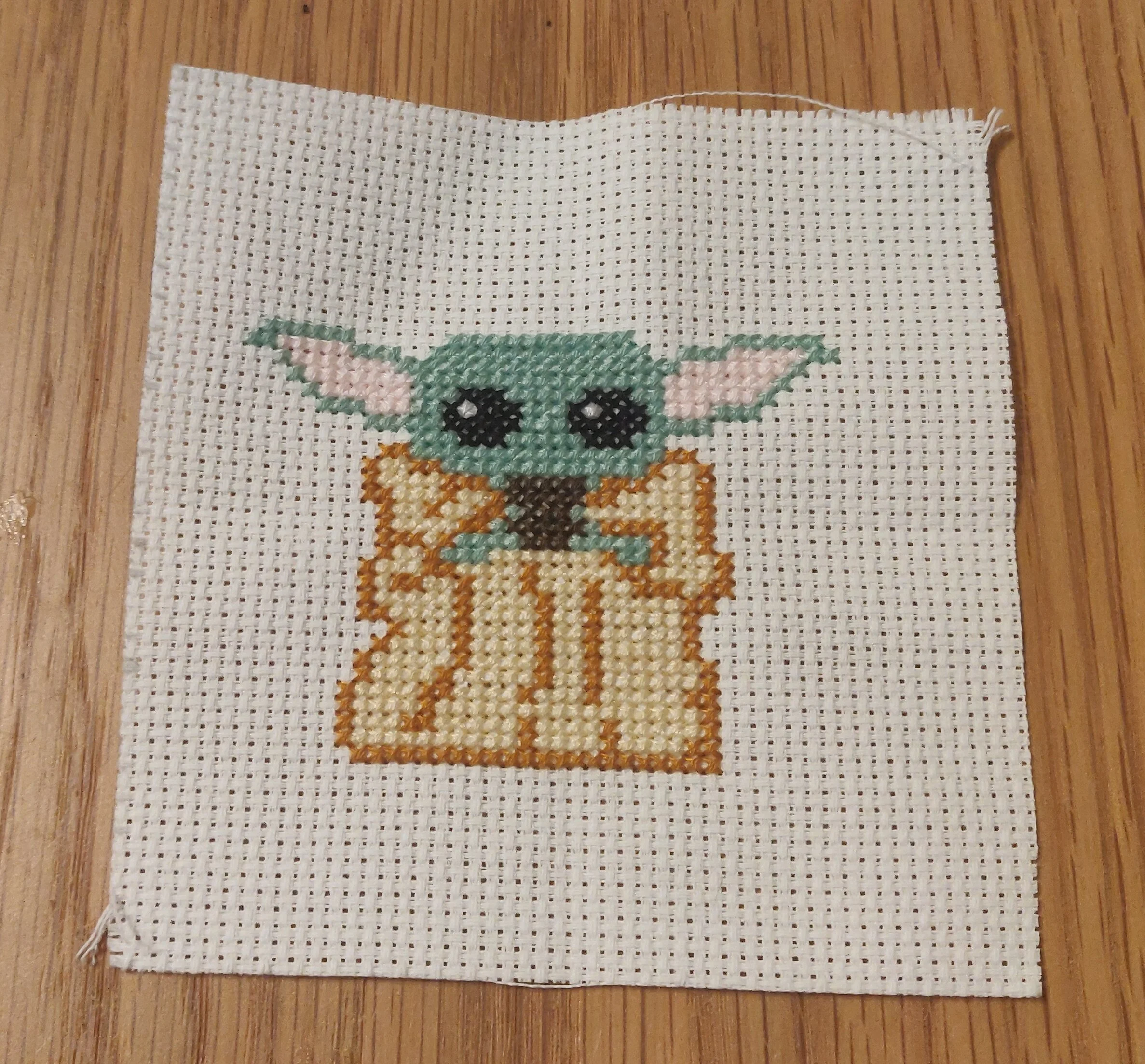 White Aida fabric with the Star Wars character Grogu cross-stitched, laid on a wood background.